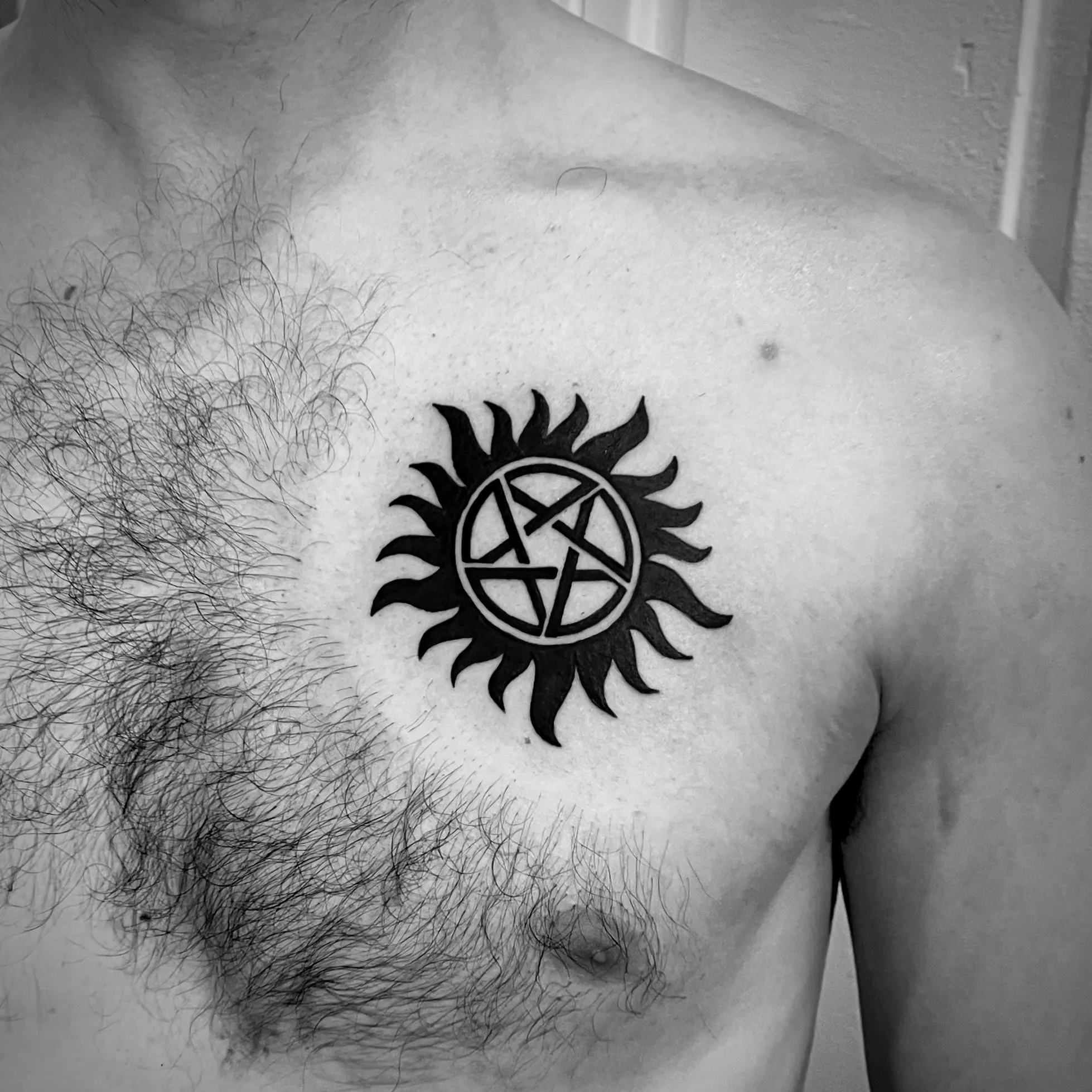 A black tattoo of a sun surrounding a pentagram with the letters A, R, S, and A inside, located on a person's upper chest near the shoulder.