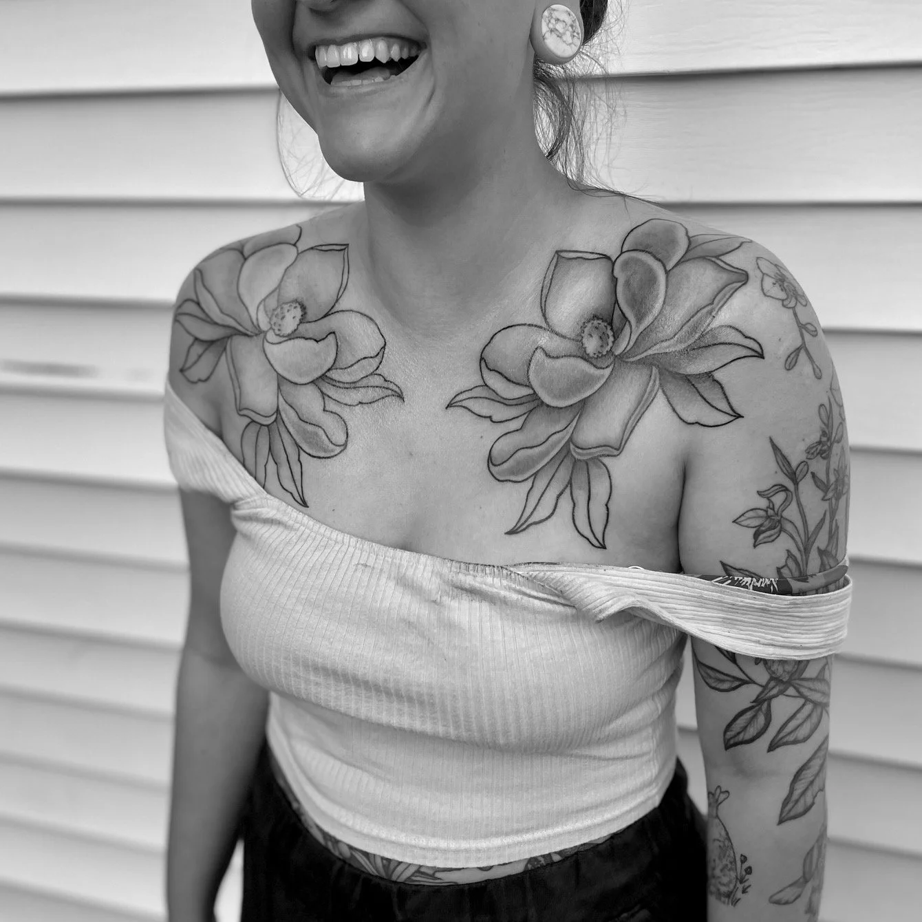 A woman with tattoos on her chest, arms, and shoulder smiling and wearing a off-shoulder top, standing in front of a horizontal siding wall.