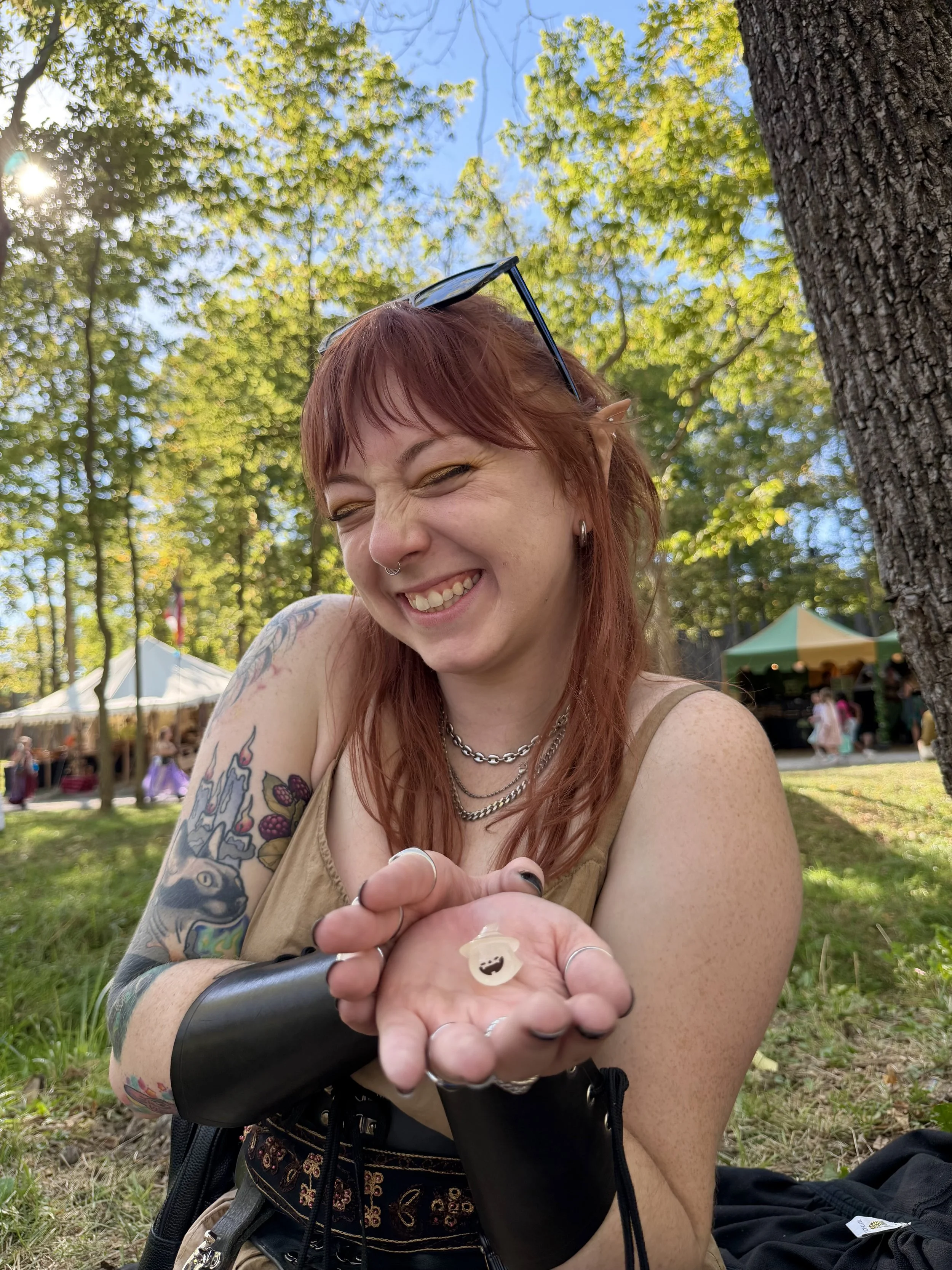 A woman with red hair, tattoos, and piercings sitting outdoors under a tree, smiling and holding small figurines or objects in her hand, with a background of tents and people at an outdoor event.