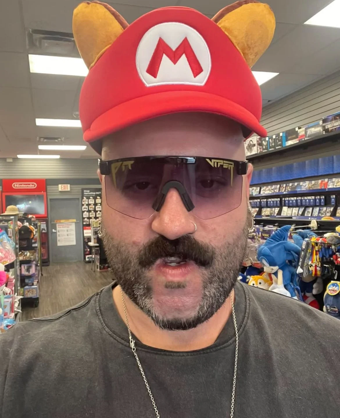 Man wearing a red Mario hat with yellow ears, black sunglasses, and a nose ring inside a store aisle.