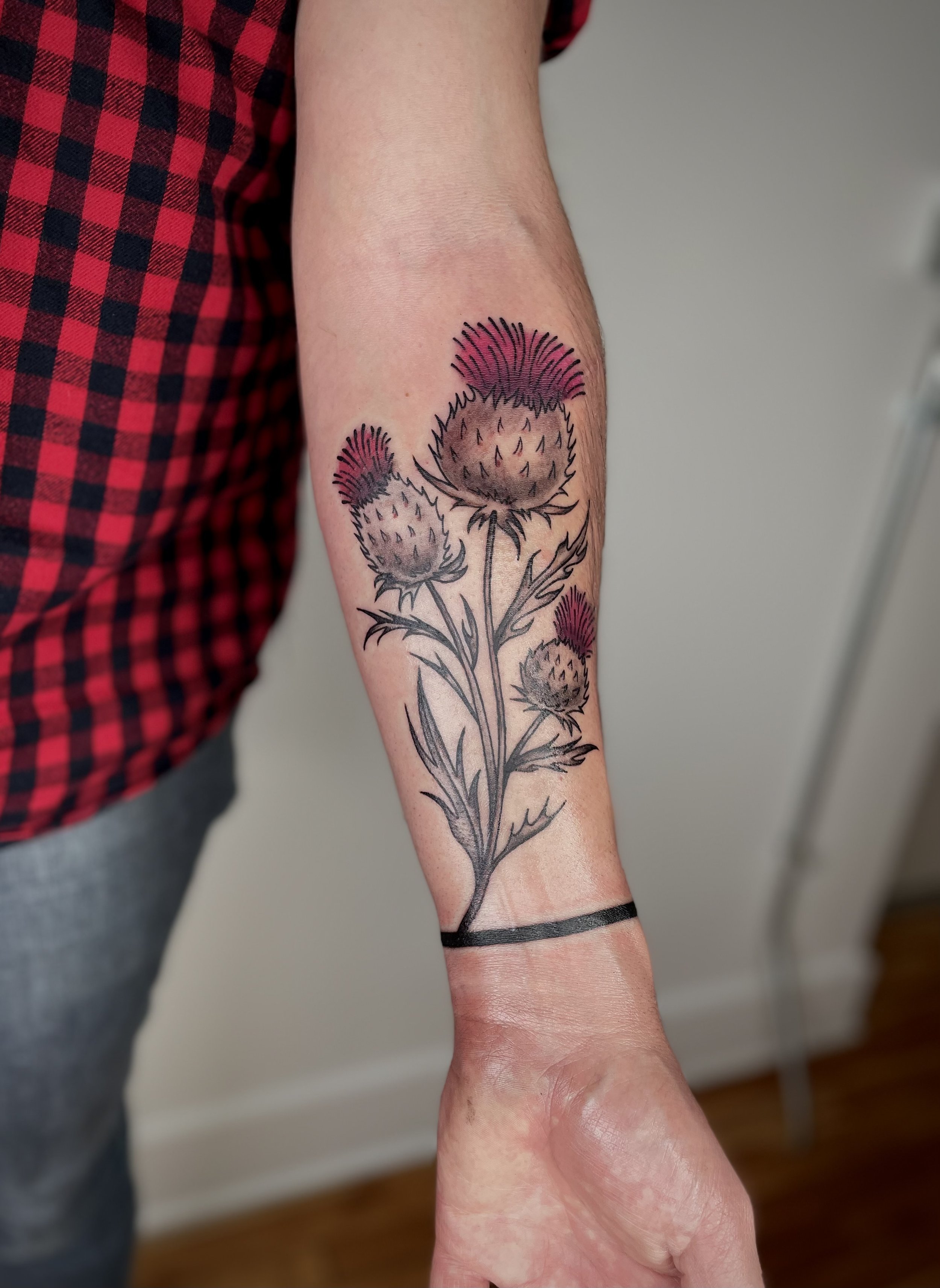 Tattoo of thistle flowers on person's forearm with black outline and pink accents, extending from wrist to mid-forearm.