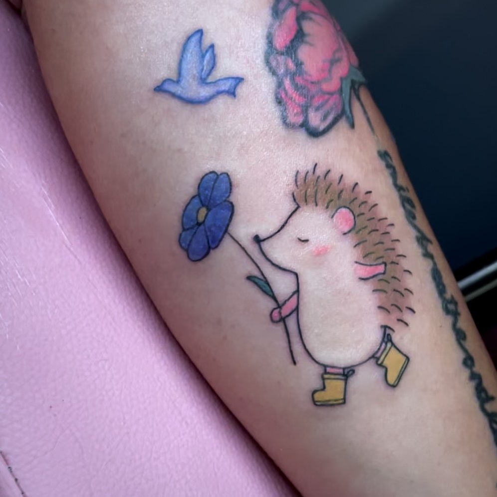 A colorful tattoo of a cute hedgehog holding a blue flower, wearing yellow rain boots, on a person's arm.