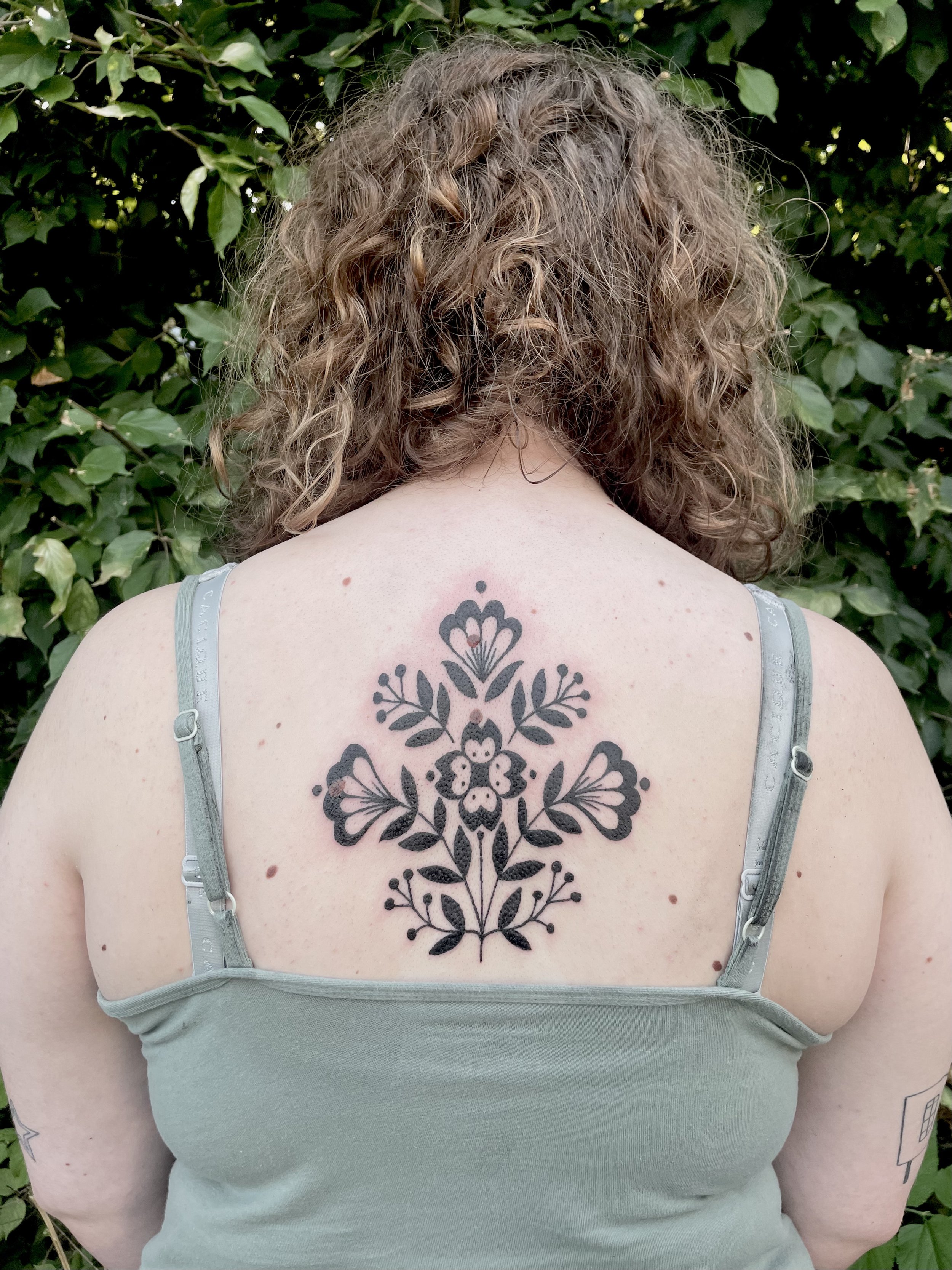 Back view of a woman with curly hair wearing a light green tank top, showing a black floral tattoo on her upper back.