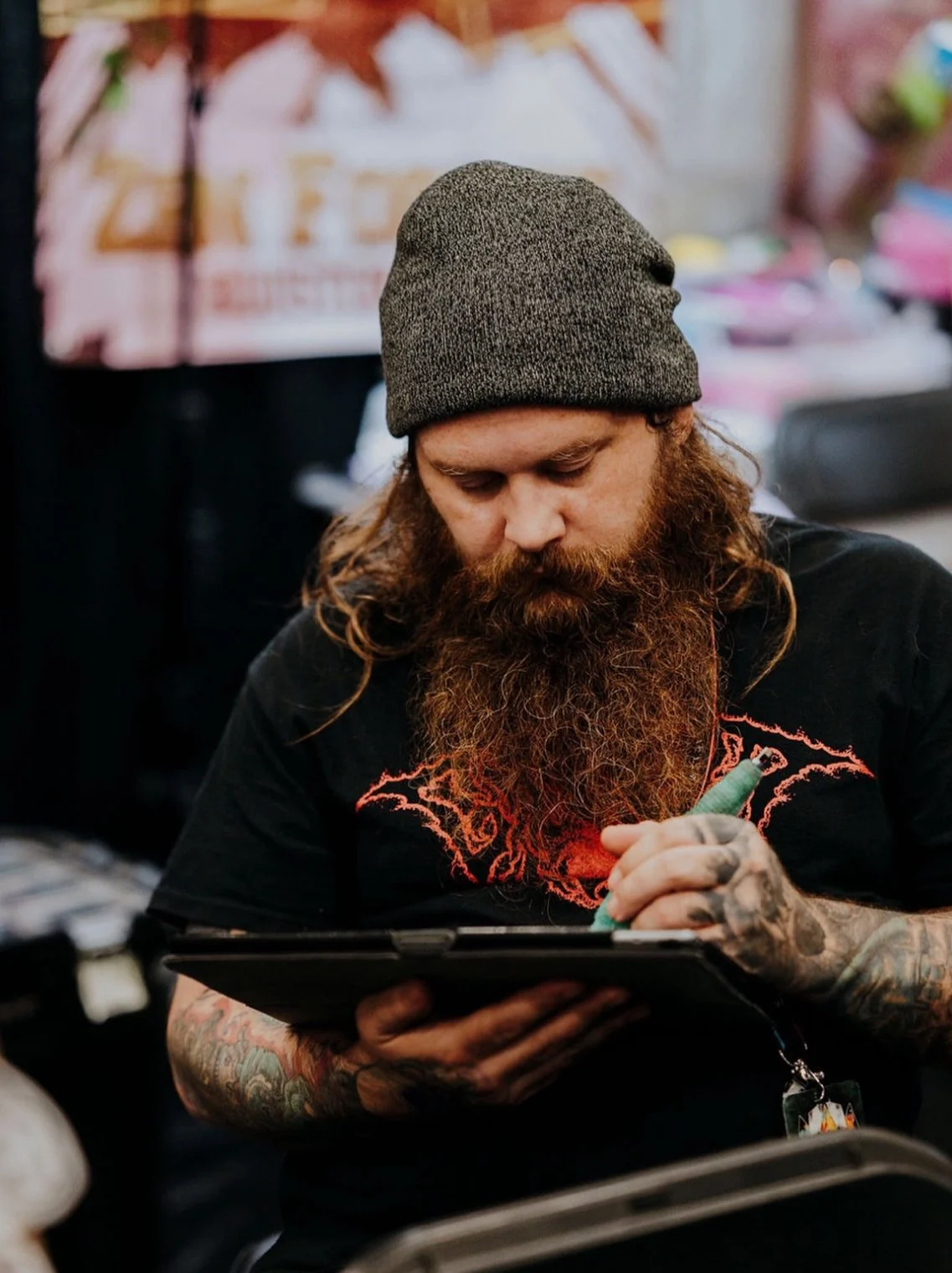 A man with a beard, tattoos, and a gray beanie is writing or drawing on a clipboard with a green marker in a setting with blurred background and a colorful poster or banner.