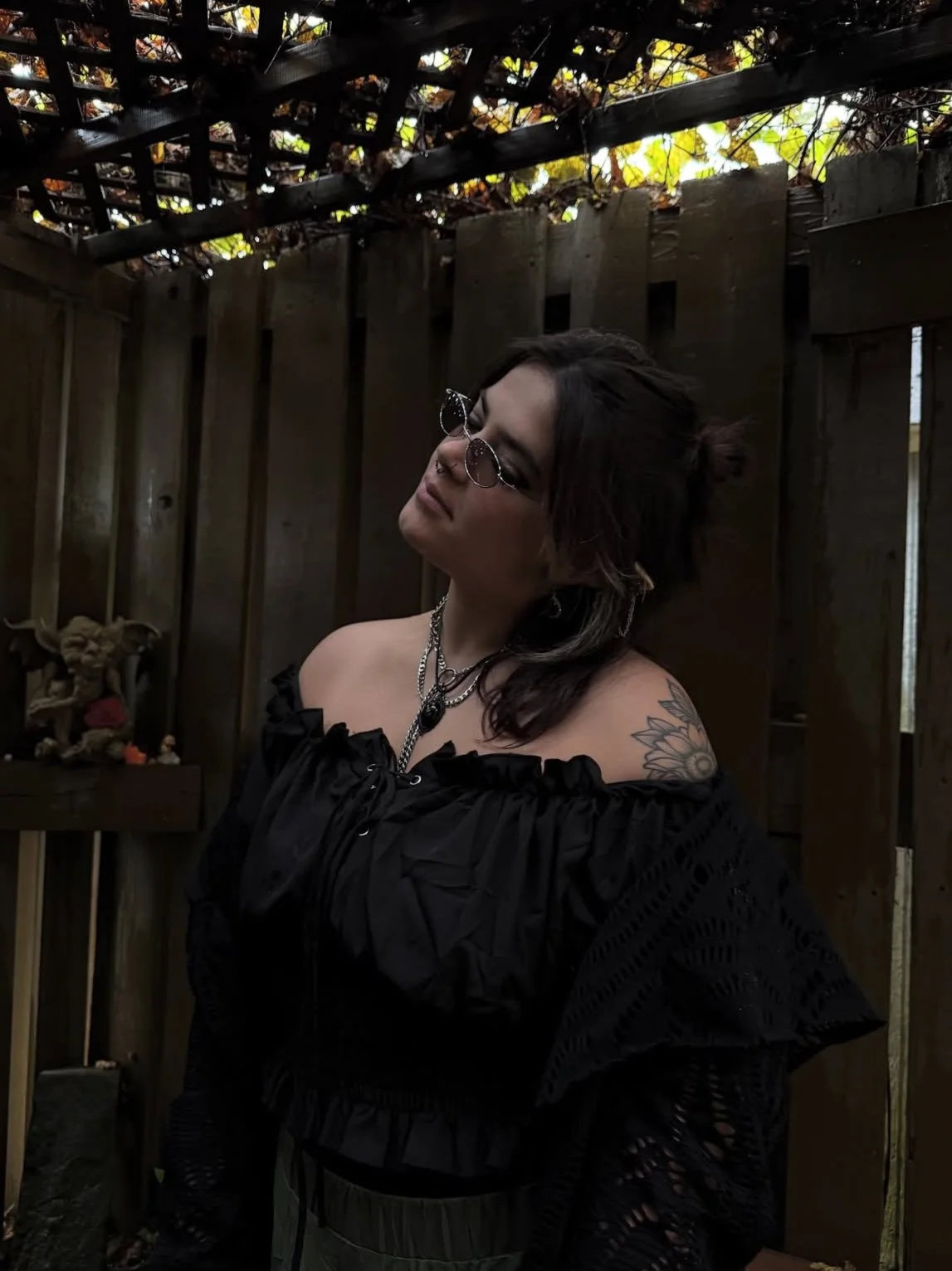 A woman with dark hair and tattoos wearing glasses, black off-shoulder top, and jewelry, standing in a wooden outdoor space with a lattice roof and autumn leaves.
