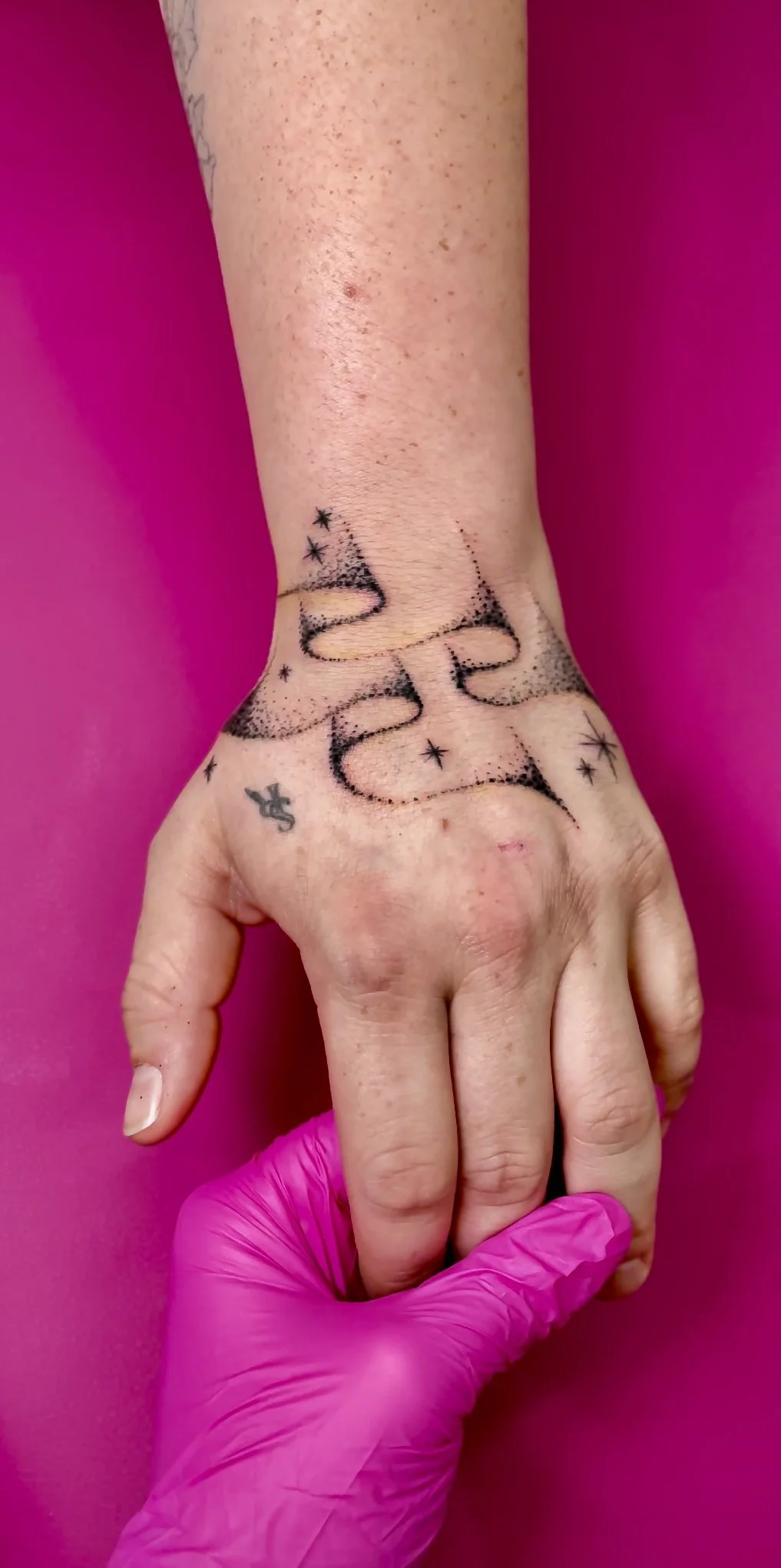 A person's hand with a black tattoo on the wrist and forearm, against a pink background, with a pink glove holding the hand's fingers.