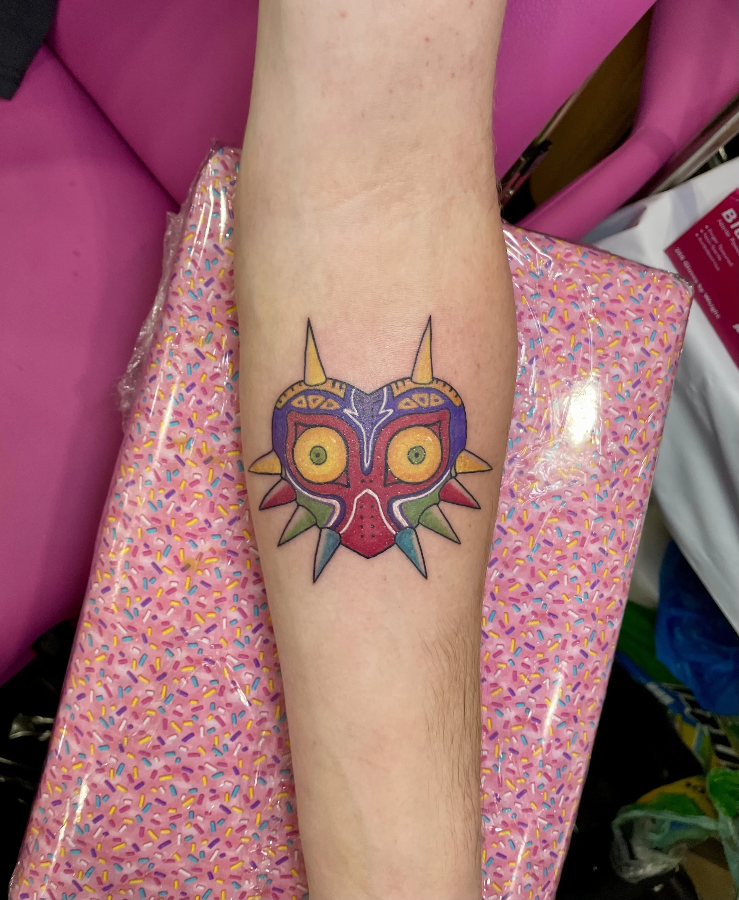 Colorful tattoo of Majora's Mask from The Legend of Zelda on a person's forearm, resting on pink, sprinkle-patterned fabric.