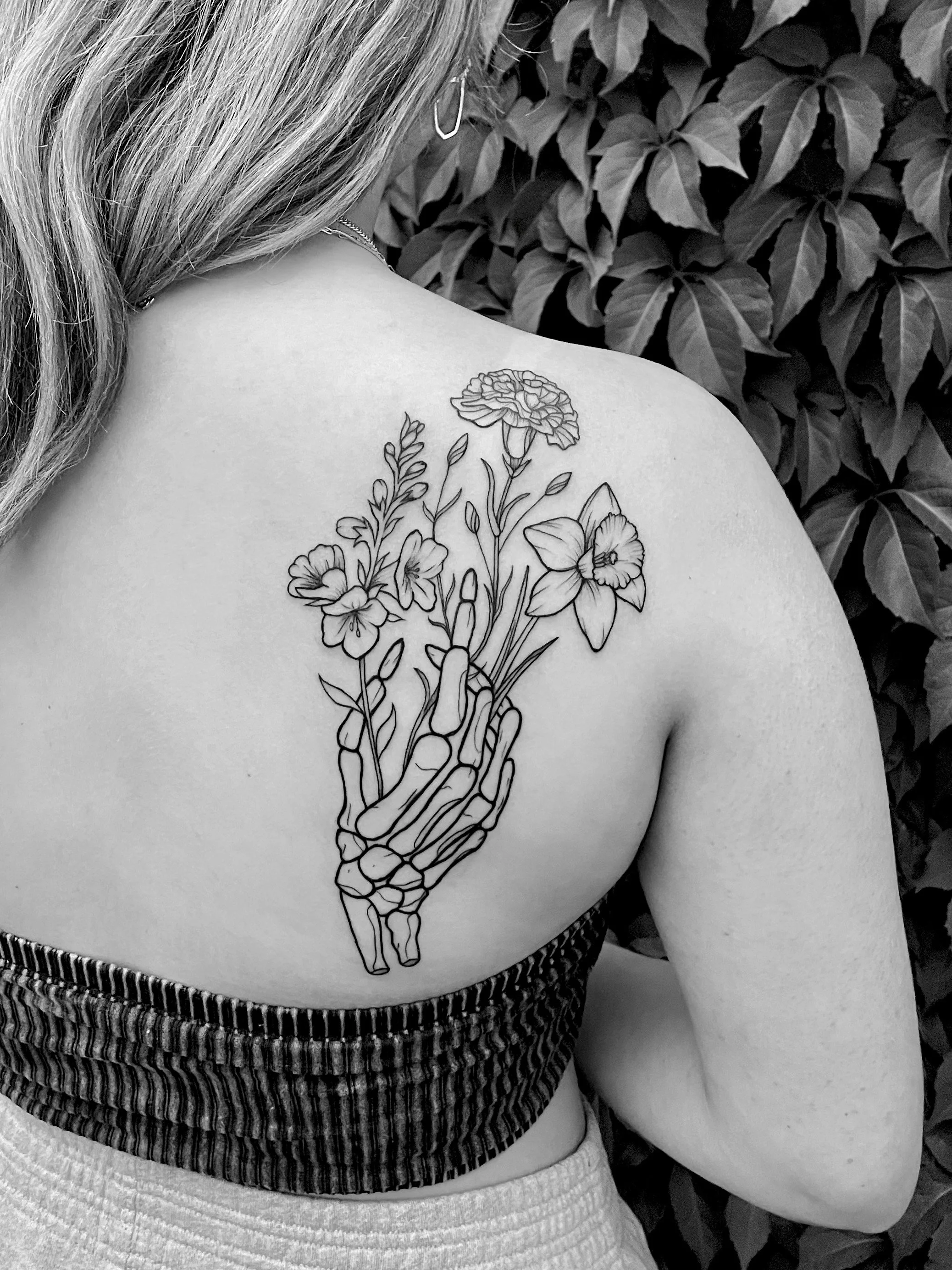 A black-and-white photo of a woman with a tattoo on her left shoulder blade of a skeletal hand holding a bouquet of flowers, with dark foliage in the background.
