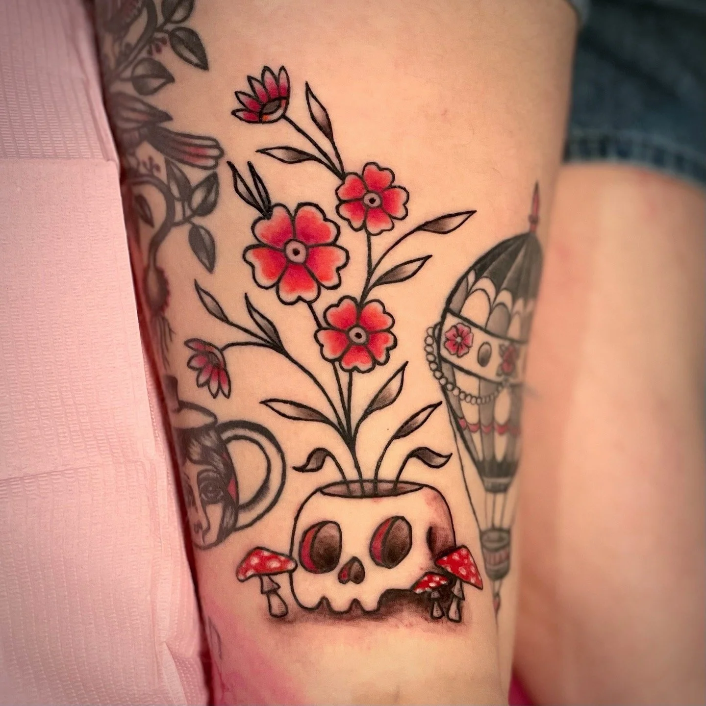 Tattoo of a skull with flowers growing out of it, mushrooms around the base, and a hot air balloon in the background.