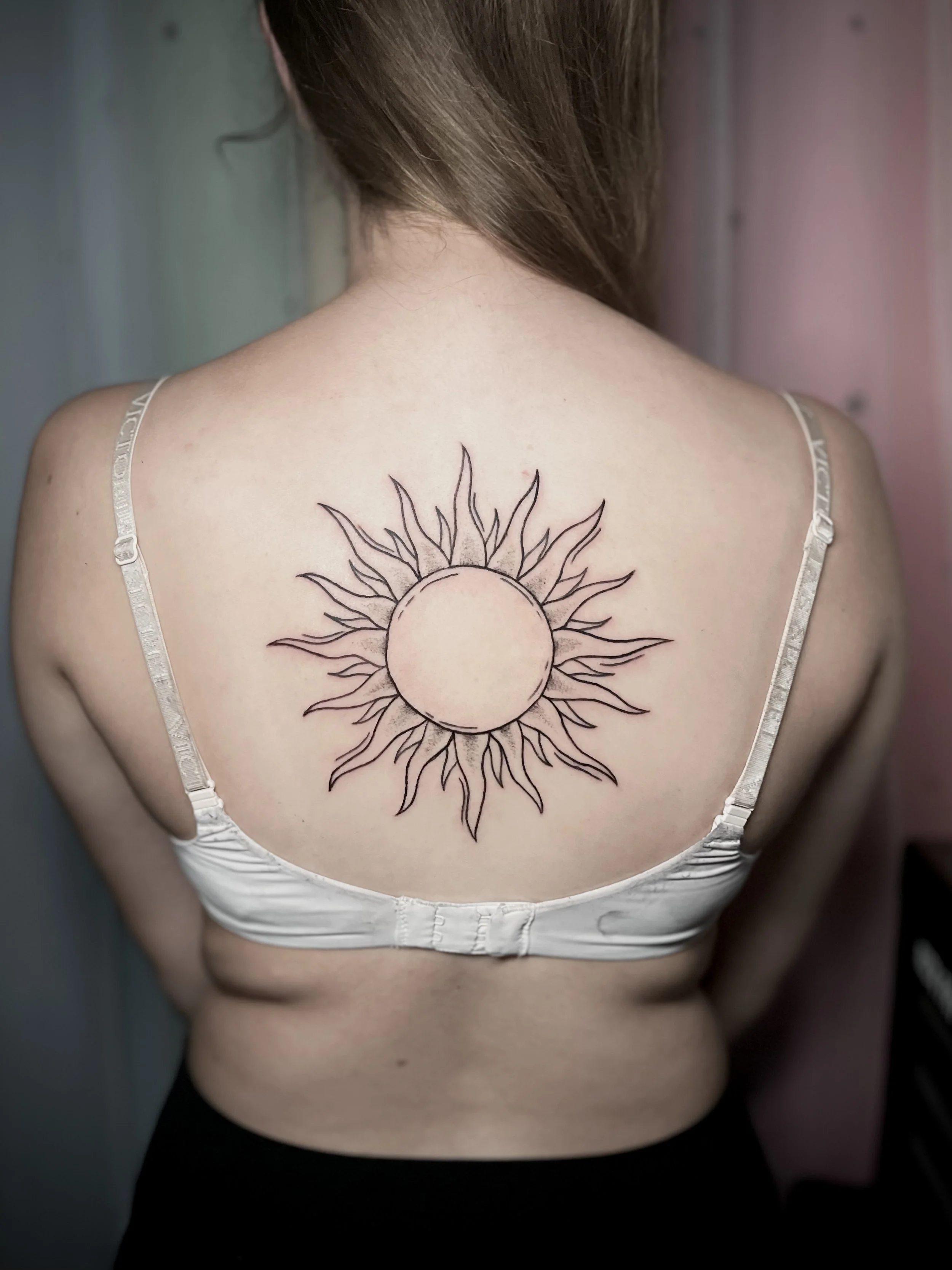 Tattoo of a circular sun with radiating wavy lines on a woman's upper back.