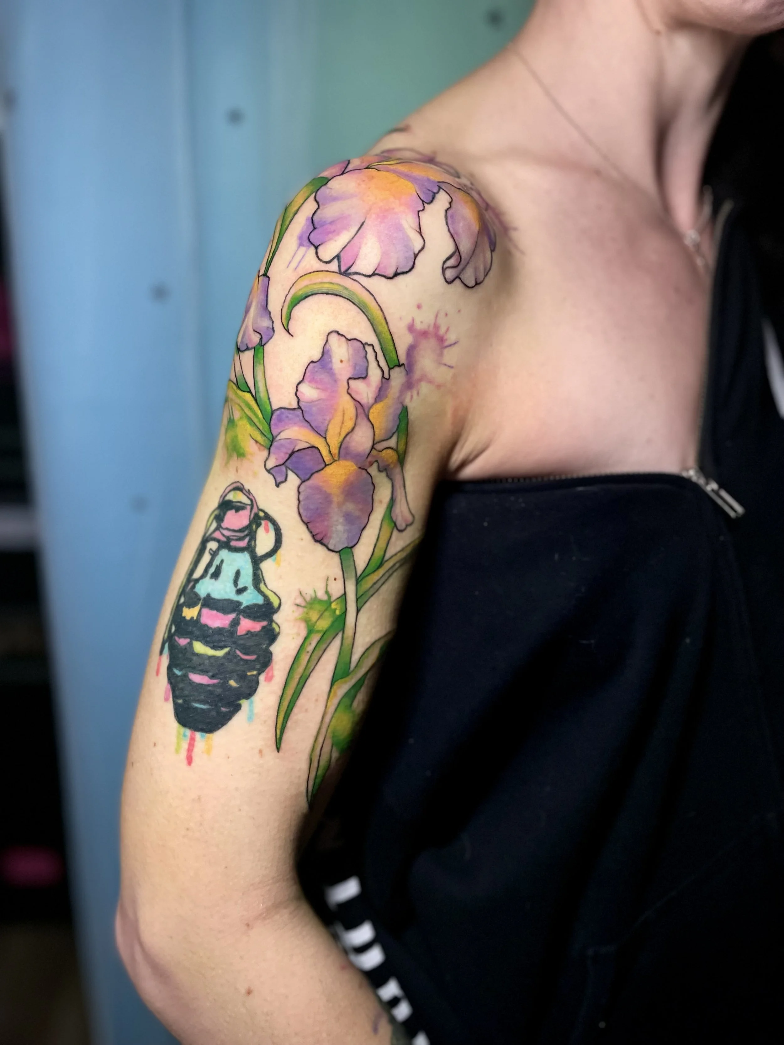 Colorful floral tattoo of irises on woman's upper arm and shoulder, with a small tattoo of a dripping iced coffee cup on her lower arm.