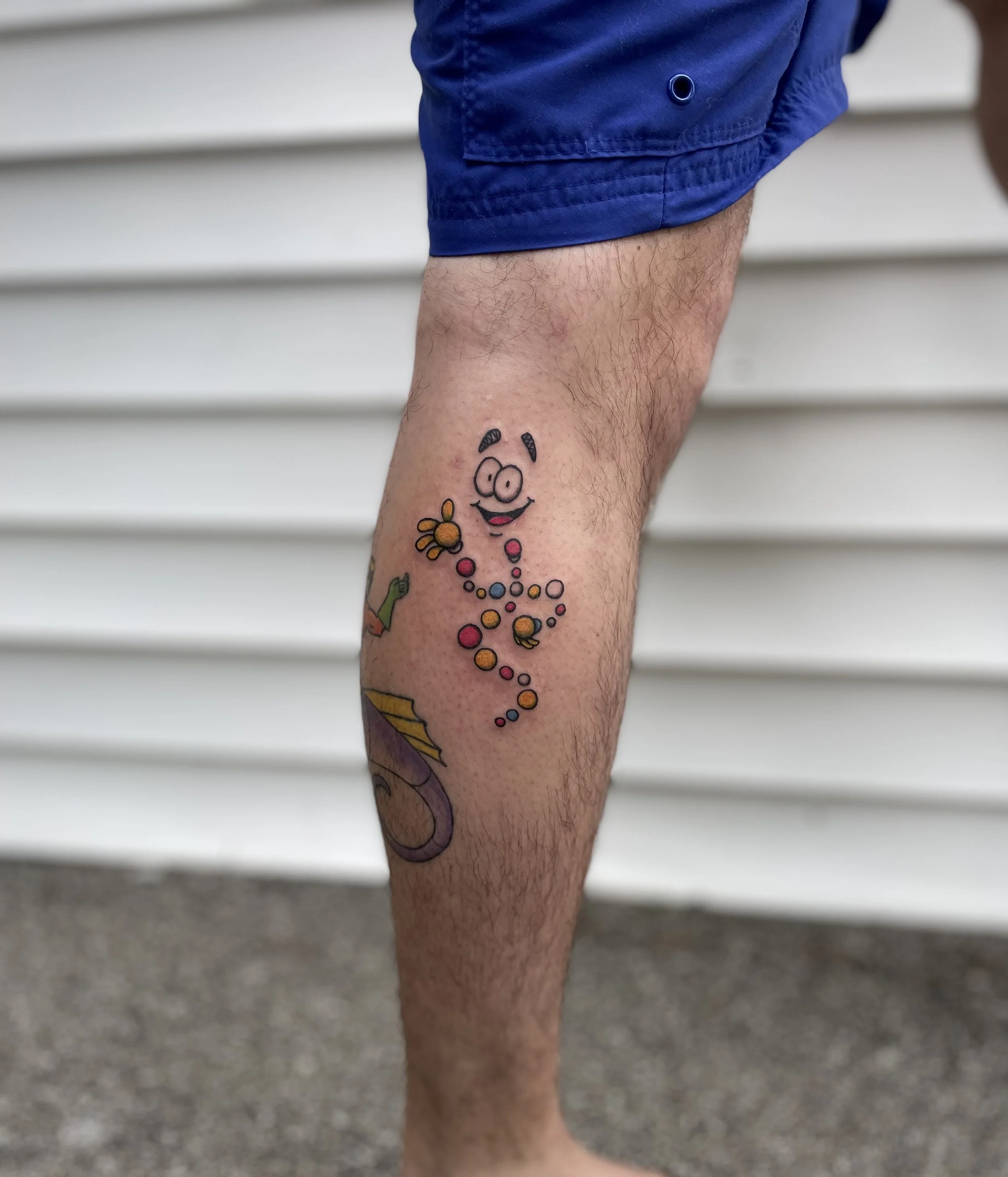 A tattoo on a person's leg depicts a cartoon smiley face with eyes, nose, and mouth, surrounded by colorful dots, with additional small images partially visible below.