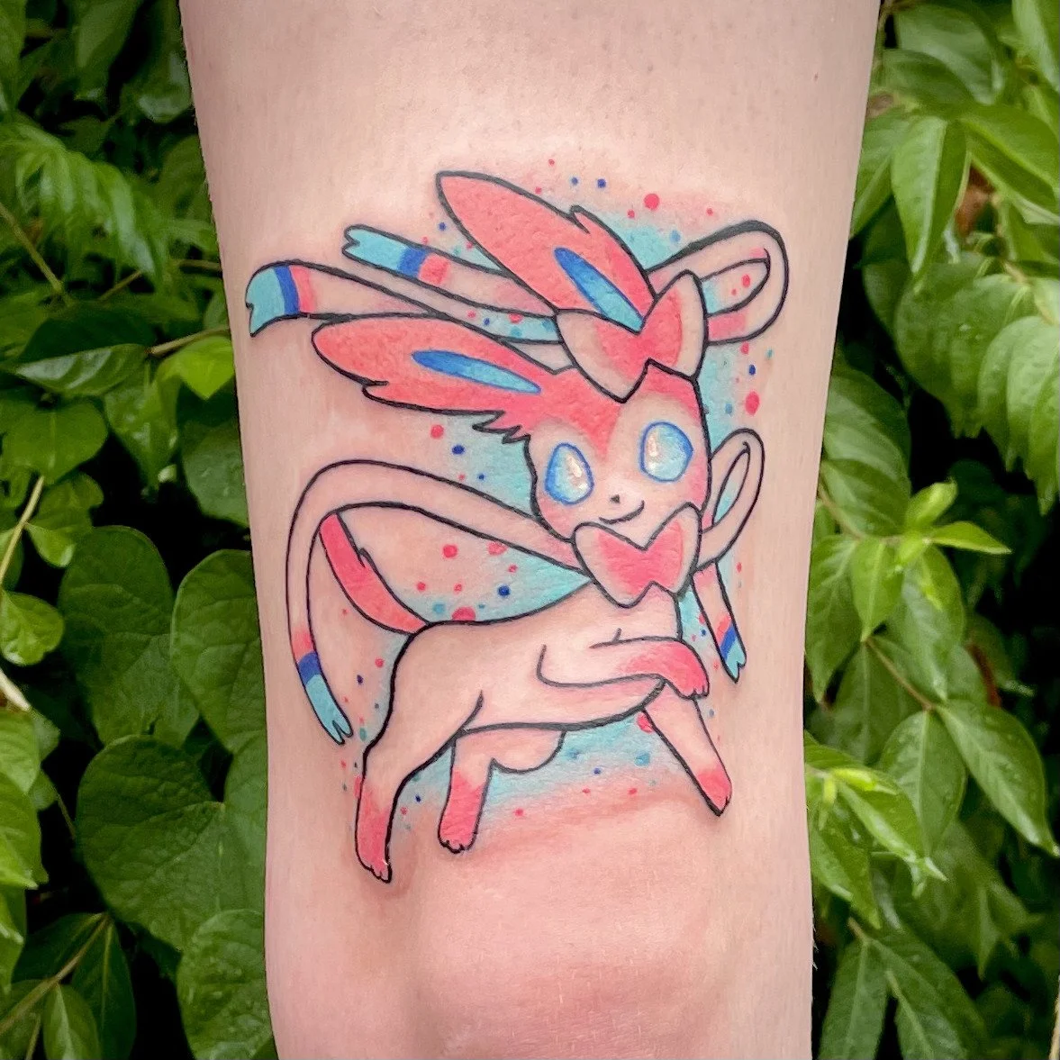 A colorful tattoo of a mythical creature with a feline body, large wings, and a skull-like face with blue eyes, surrounded by blue and red splashes, on a person's arm.