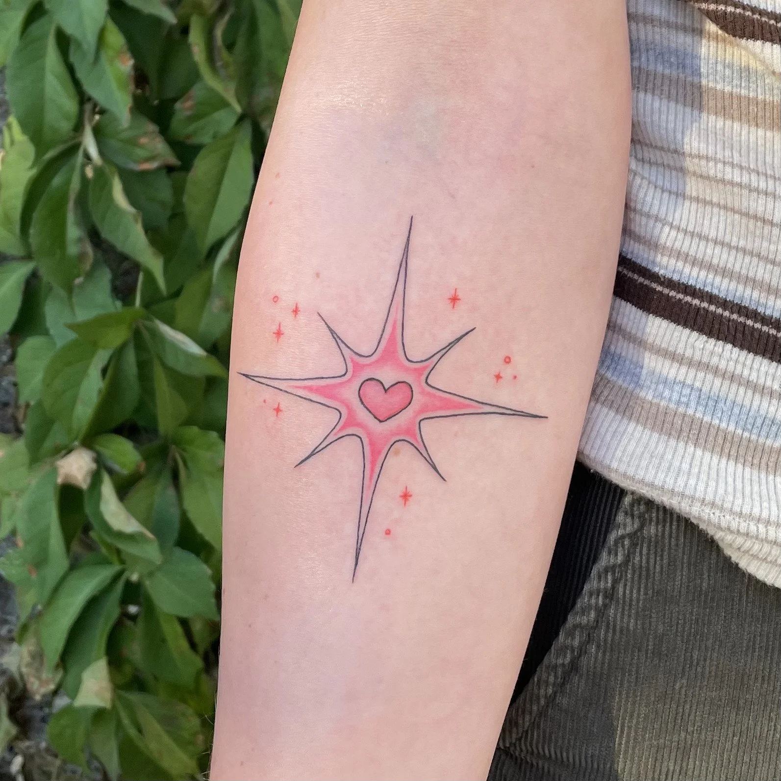Tattoo of a starburst design with a pink heart in the center, surrounded by small red stars and dots on a person's arm.