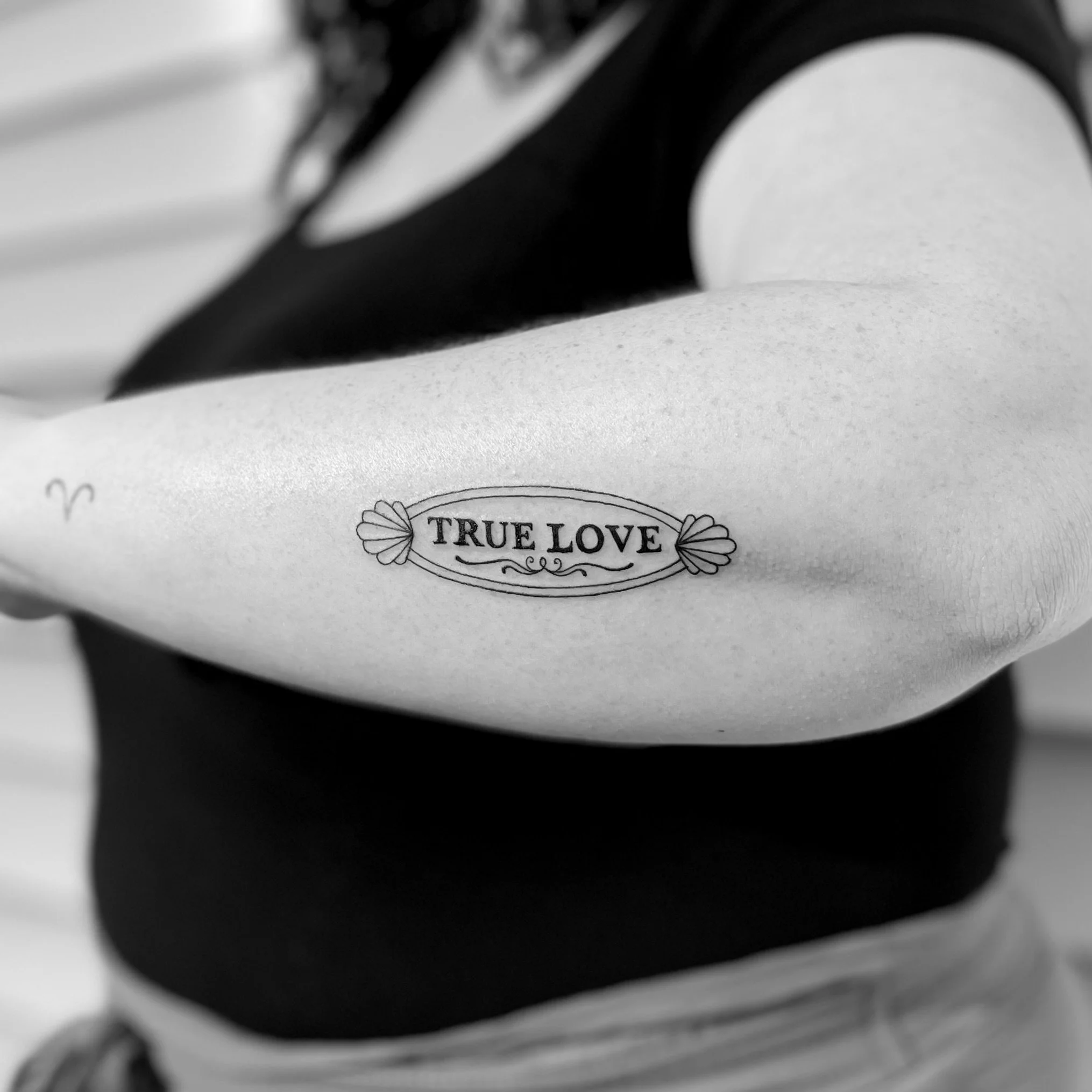 Tattoo on person's arm that says 'TRUE LOVE' inside an oval with decorative elements, in black and white.