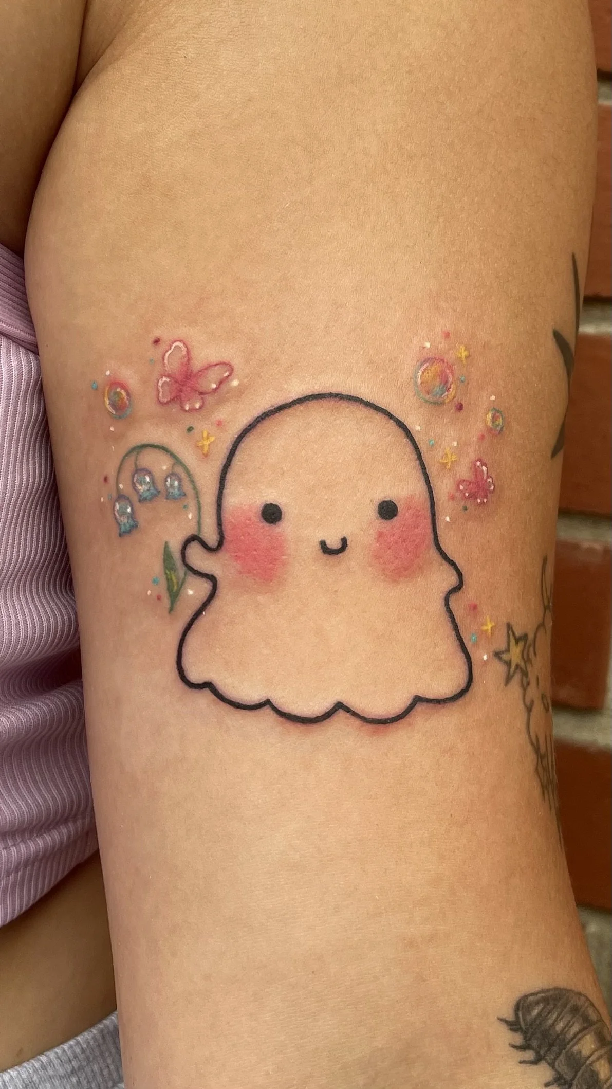 A colorful tattoo of a cute, smiling ghost with blushing cheeks, surrounded by small decorative elements like butterflies, stars, and bubbles.