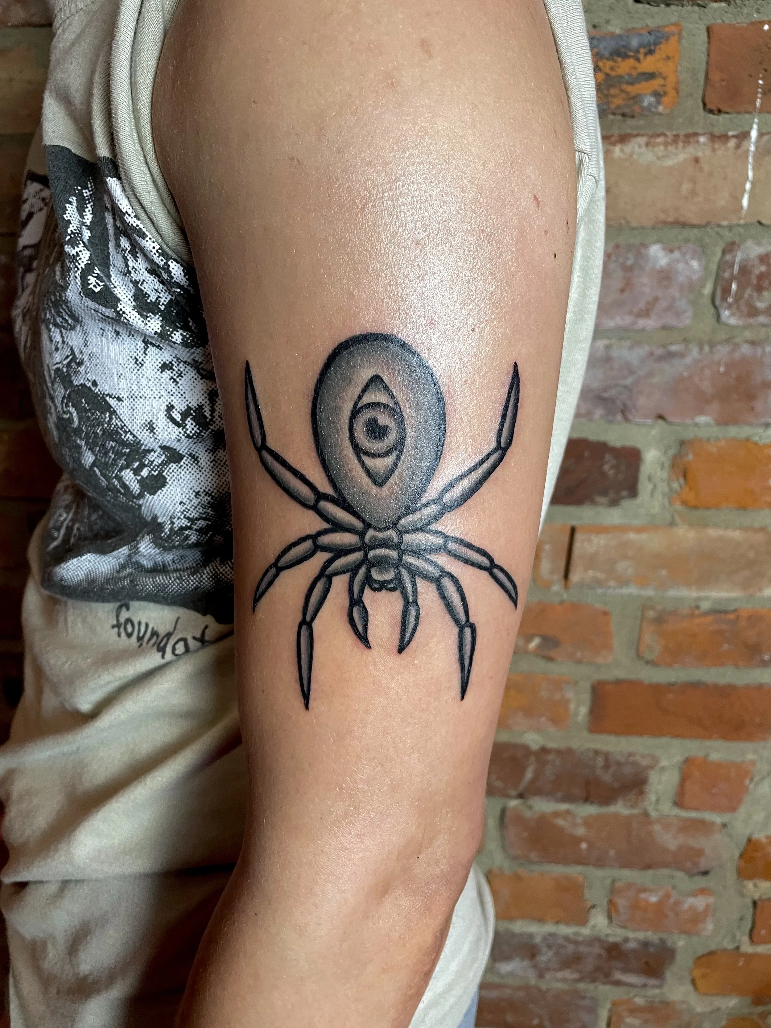 Tattoo of a spider with an eye on its abdomen on a person's upper arm, with a brick wall in the background.