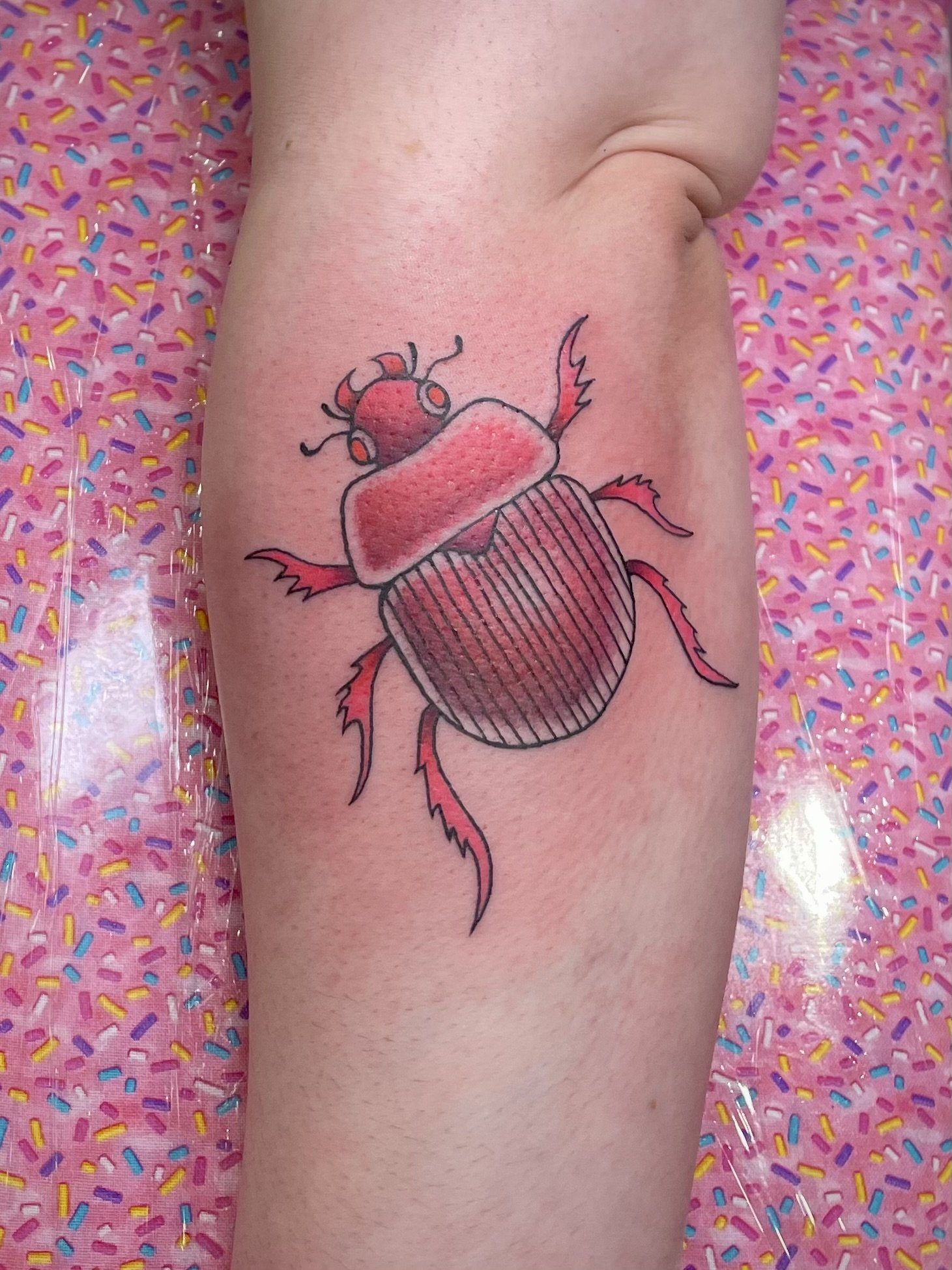 A tattoo of a beetle on a person's leg, with pink and black shading on the beetle and a pink background with colorful sprinkles.