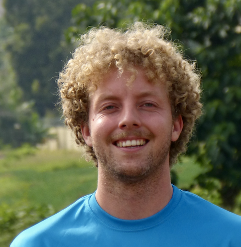 A smiling man with curly blonde hair wearing a blue shirt outdoors