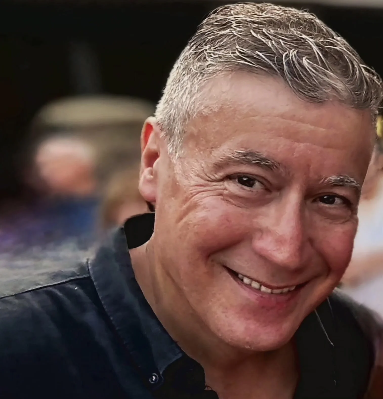 Close-up of smiling middle-aged man with short gray hair, wearing a dark shirt, at a social gathering.
