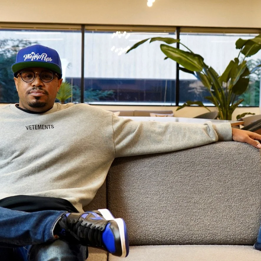 Man sitting on a couch, wearing glasses, a blue and green cap, a beige sweatshirt, and black and blue sneakers, in a modern indoor setting with large windows and a potted plant in the background.