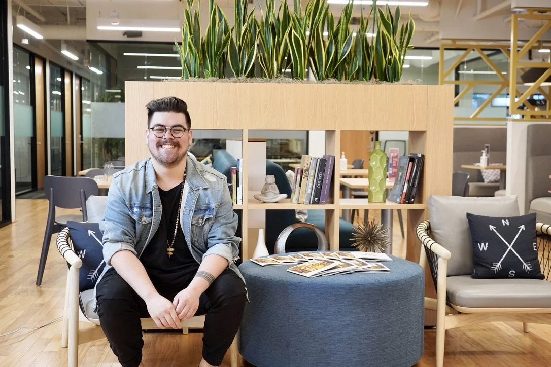 A smiling man with glasses, wearing a jean jacket and black clothing, sitting on a modern chair in a well-lit indoor space with shelves, plants, and decorative items in the background.