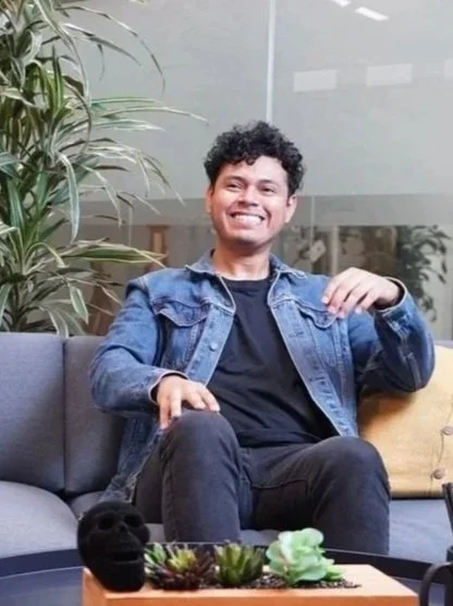 A young man with curly hair smiling, sitting on a dark gray couch with a yellow pillow, next to a black dog, in a modern indoor space with plants.