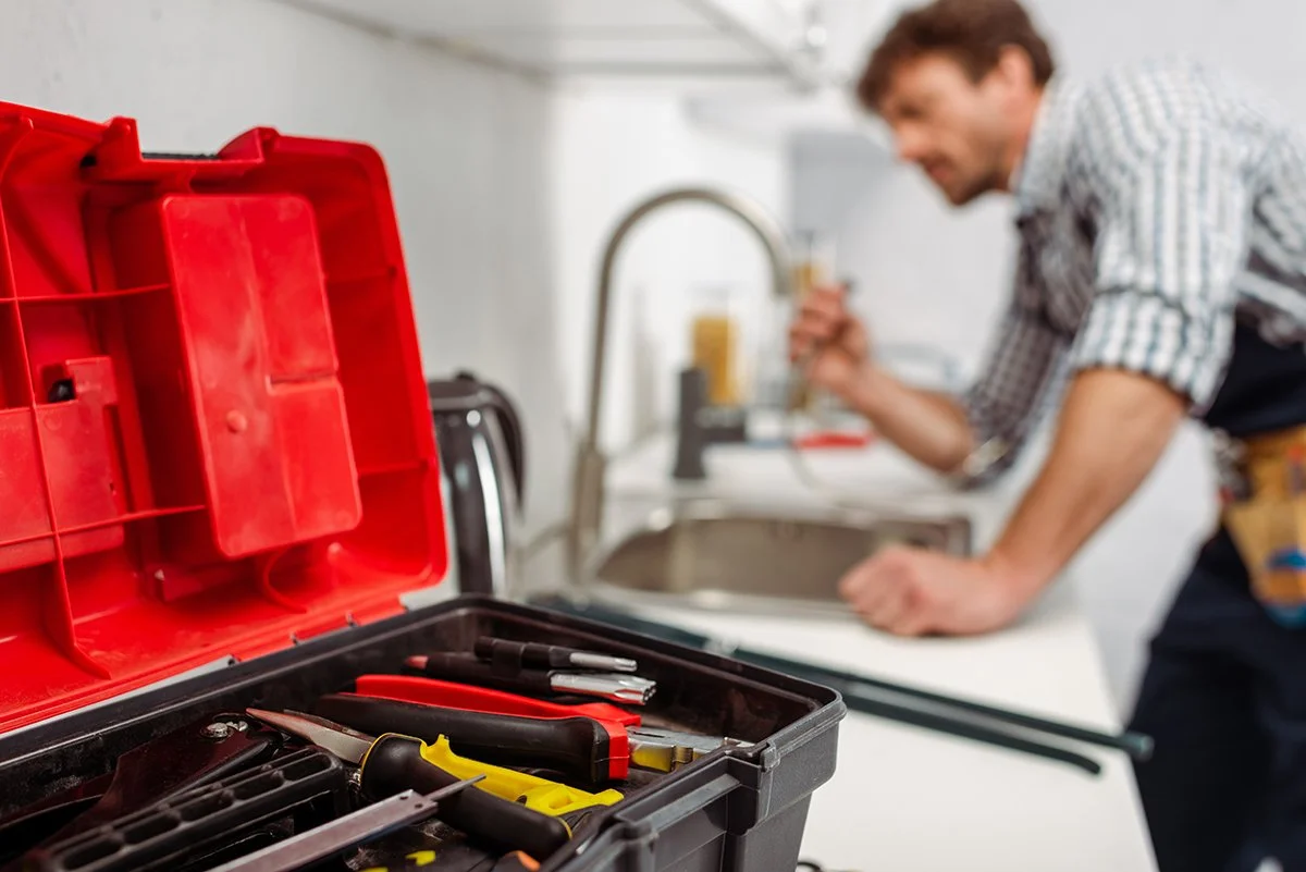 Open toolbox with various hand tools on a work surface, with a blurry man in the background fixing or installing something at a sink.