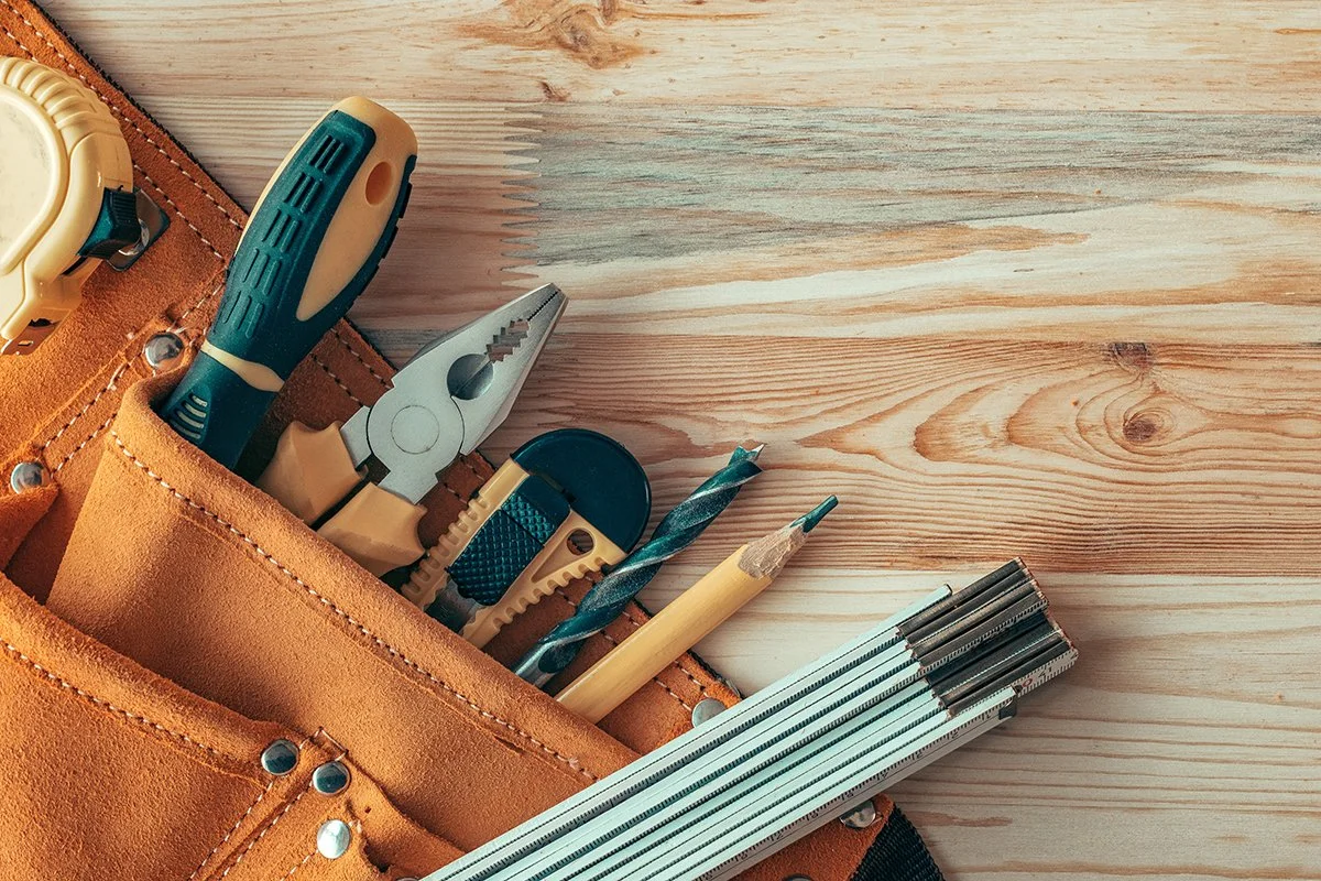 A leather tool belt on a wooden surface with various tools including a screwdriver, pliers, utility knife, drill bit, and a bundle of drill bits.
