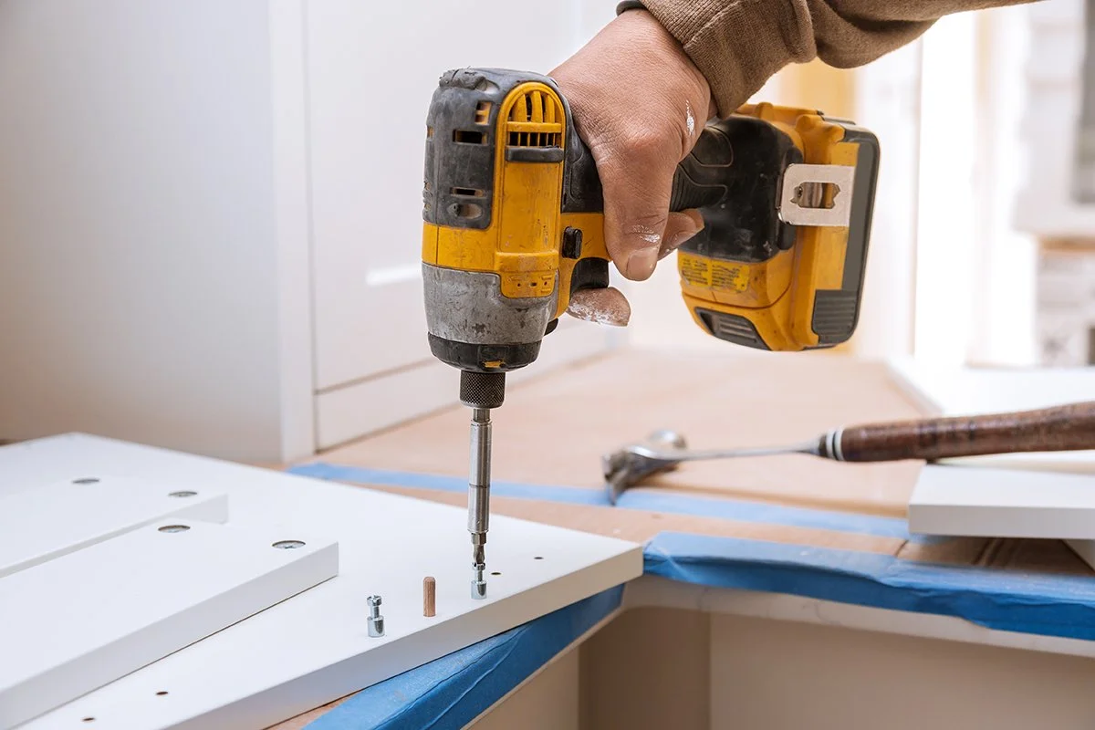 A person using a yellow cordless drill to assemble white furniture on a countertop covered with blue painter's tape.