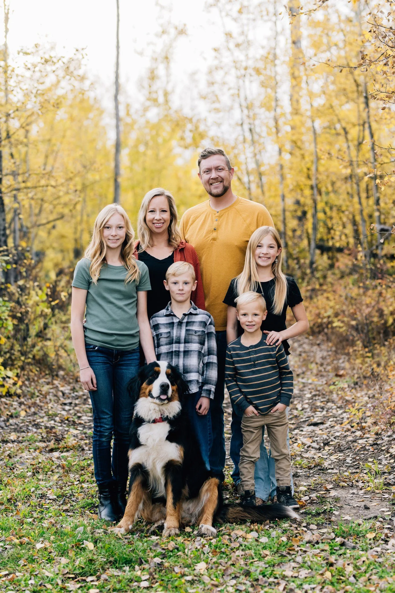 A smiling family of seven, including a dog, standing outdoors on a forest path with yellow fall foliage in the background.