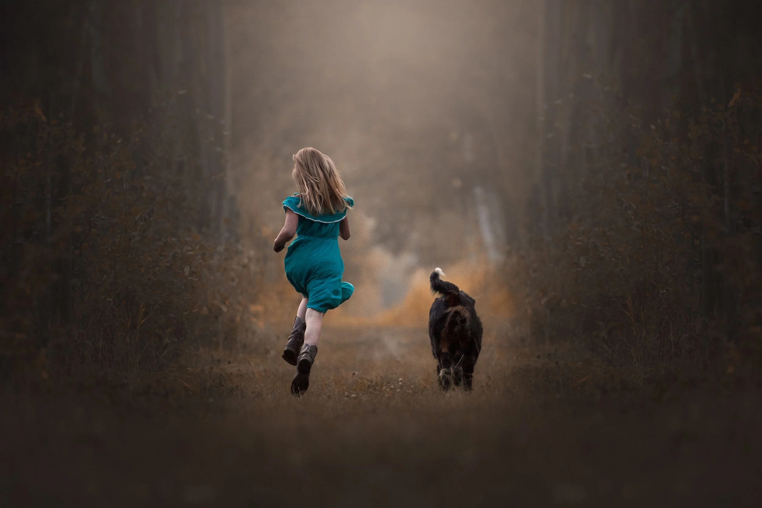 A young girl in a teal dress running with a black dog down a woodland path during late afternoon or early evening.
