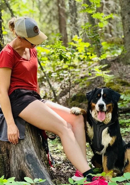 Woman sitting on a tree stump in a forest, wearing a red shirt, black shorts, and a baseball cap, with a black, white, and brown dog sitting beside her.