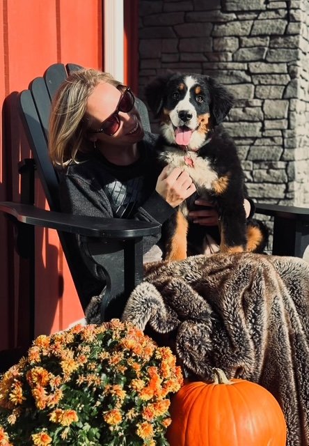 Woman sitting in a black Adirondack chair outside, holding a puppy with black, white, and brown fur, next to a large orange pumpkin and a potted plant with orange flowers, in front of a stone wall.