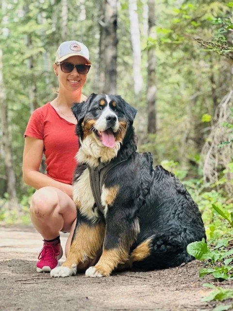 A woman in a red shirt, shorts, sun hat, and sunglasses kneels next to a large, black and tan Bernese Mountain Dog sitting on a trail in a forest.