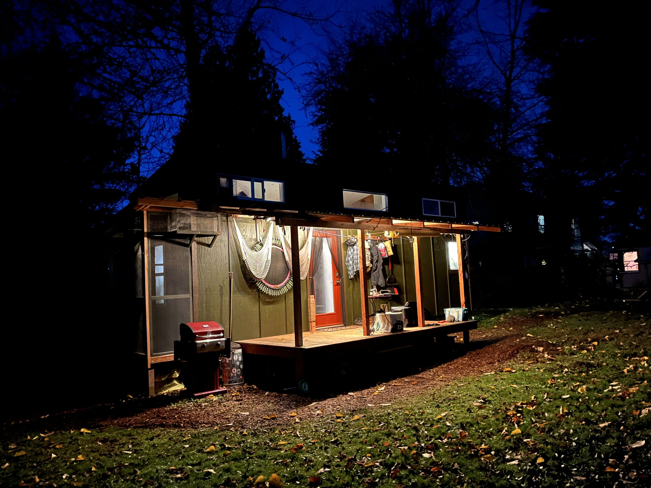 A small, partially constructed house or shed with a porch outside, illuminated at night. There are hammocks hanging on the porch, various tools and objects, and a grill on the ground nearby. The background shows a dark, wooded area with tall trees against the night sky.
