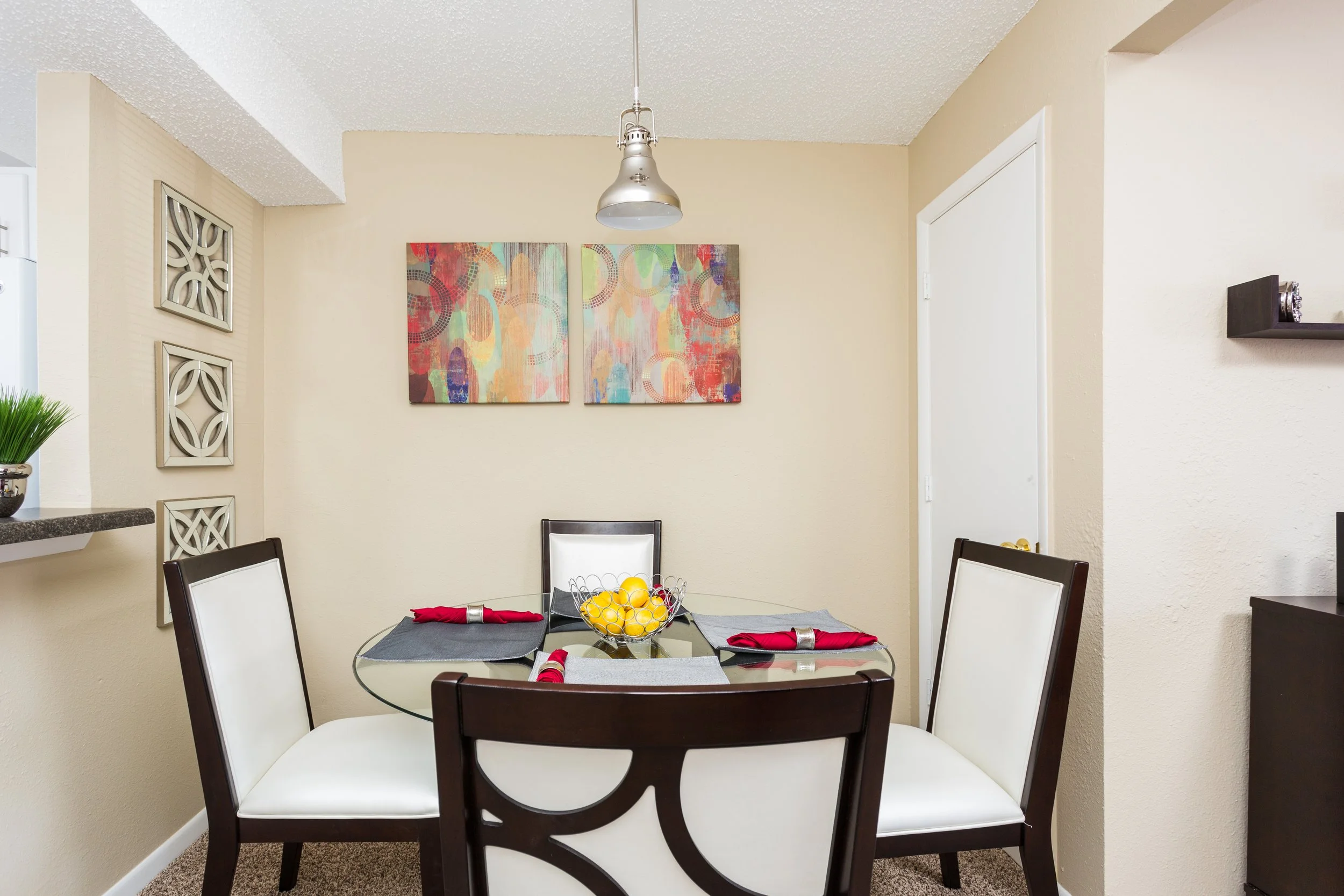 Dining room with round glass table, four white chairs with dark wood frames, yellow lemons in a wire basket, four gray placemats with red napkins, abstract colorful painting on beige wall, silver pendant light, small black shelf, decorative wall art, beige walls, carpeted floor.