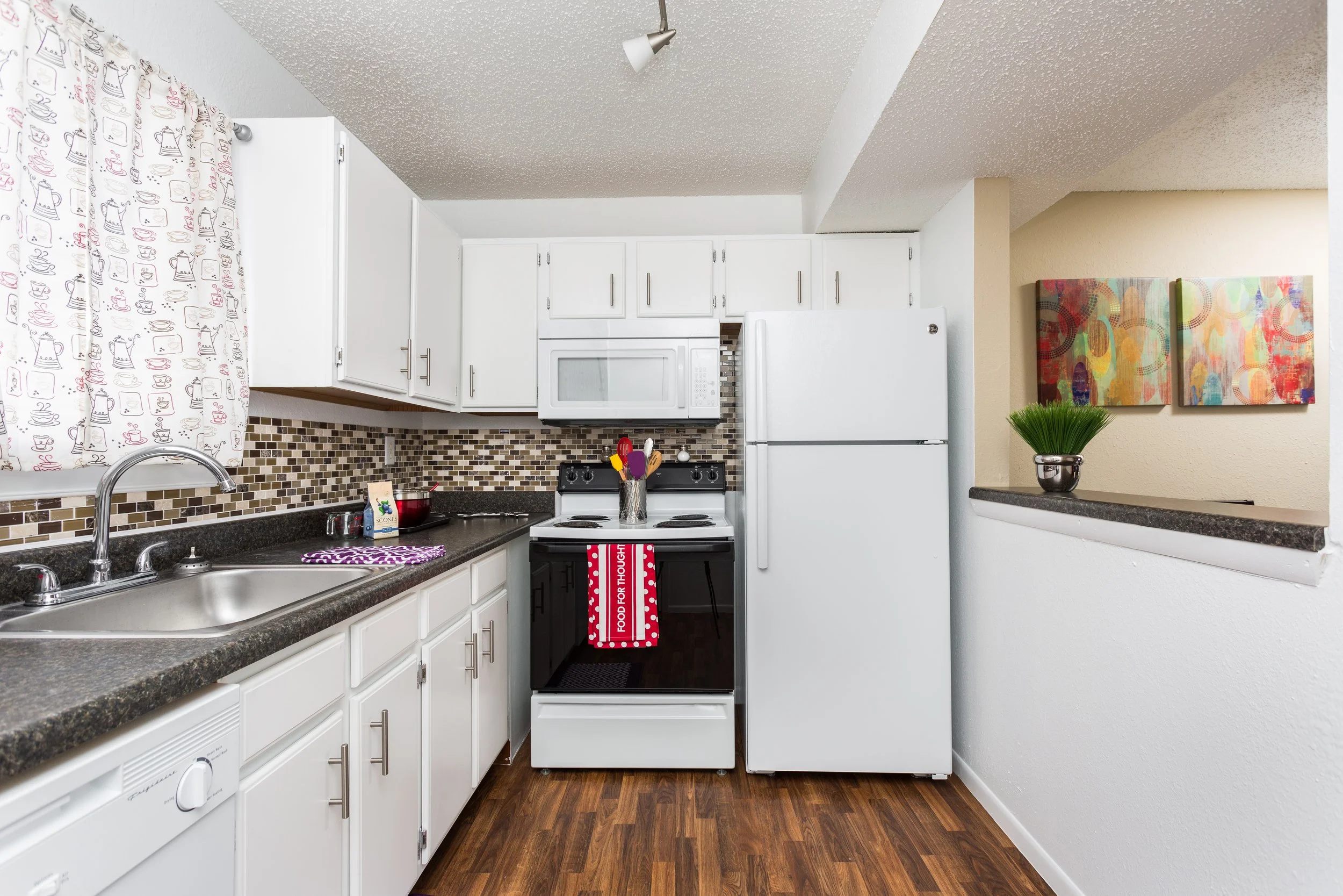 A kitchen with white cabinets, a black countertop, a white refrigerator, a stove with a towel hanging from it, and a microwave. There is a sink with a curtain with teacup and kettle patterns, and a window with curtains. The wall has a mosaic tile backsplash, and there are two colorful paintings and a green plant on a half wall.