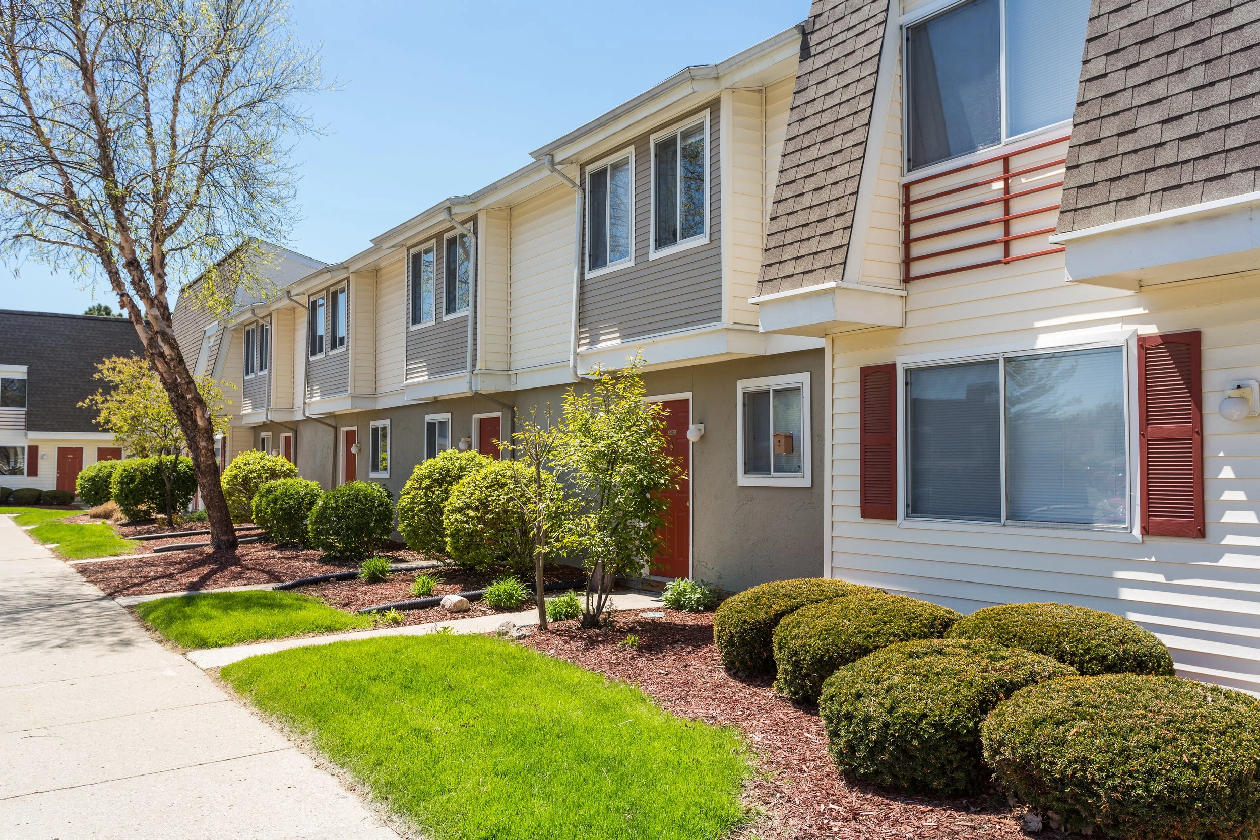 A row of townhouses with well-maintained lawn and shrubs, with a concrete sidewalk on the left, under a clear blue sky.