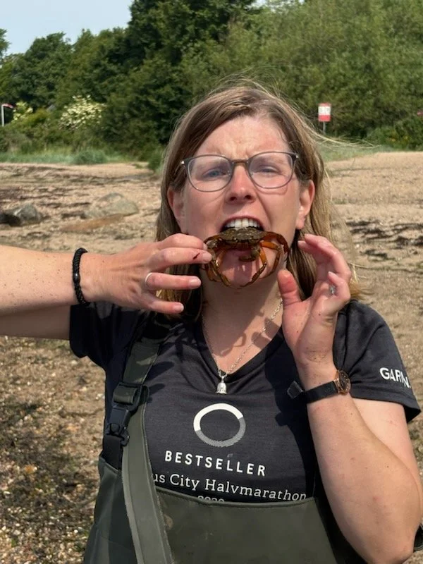 Kvinde med briller holder en krab foran munden, udendørs med træer i baggrunden.