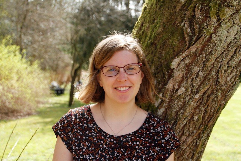 En smilende kvinde med brunt hår og briller står ved siden af et træ i en have eller park.