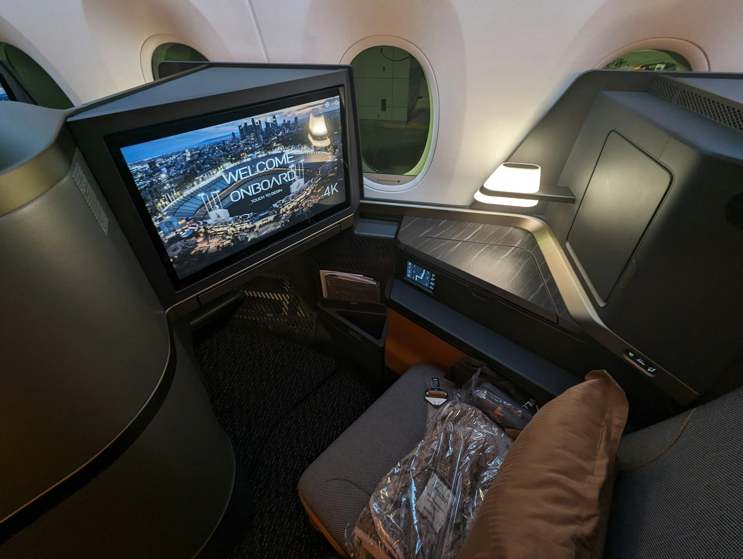 View of an airplane passenger seat with a large screen displaying a city skyline and the words 'Welcome Onboard'. The seat has a pillow, a small table, and various amenities, with windows showing the aircraft's interior structure.