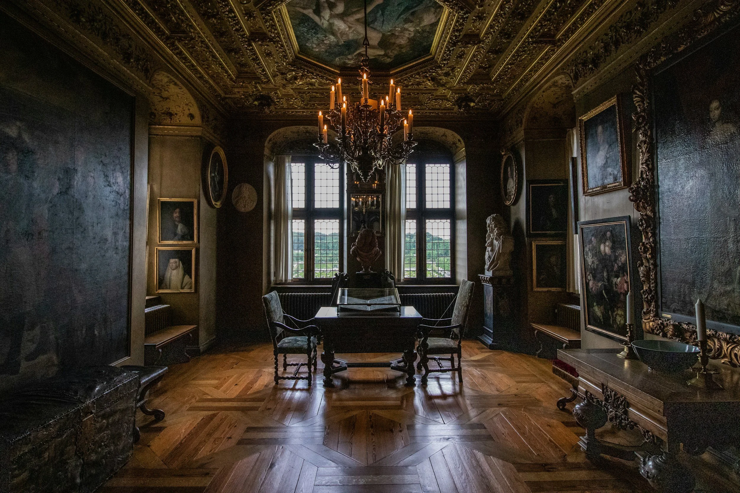 An elegant, historic room with dark wooden walls and ornate gold-framed portraits. In the center, a wooden table with a large open book and two upholstered chairs. A chandelier with lit candles hangs from a decorated ceiling, and the room is illuminated by large windows with white curtains, revealing a scenic view outside.