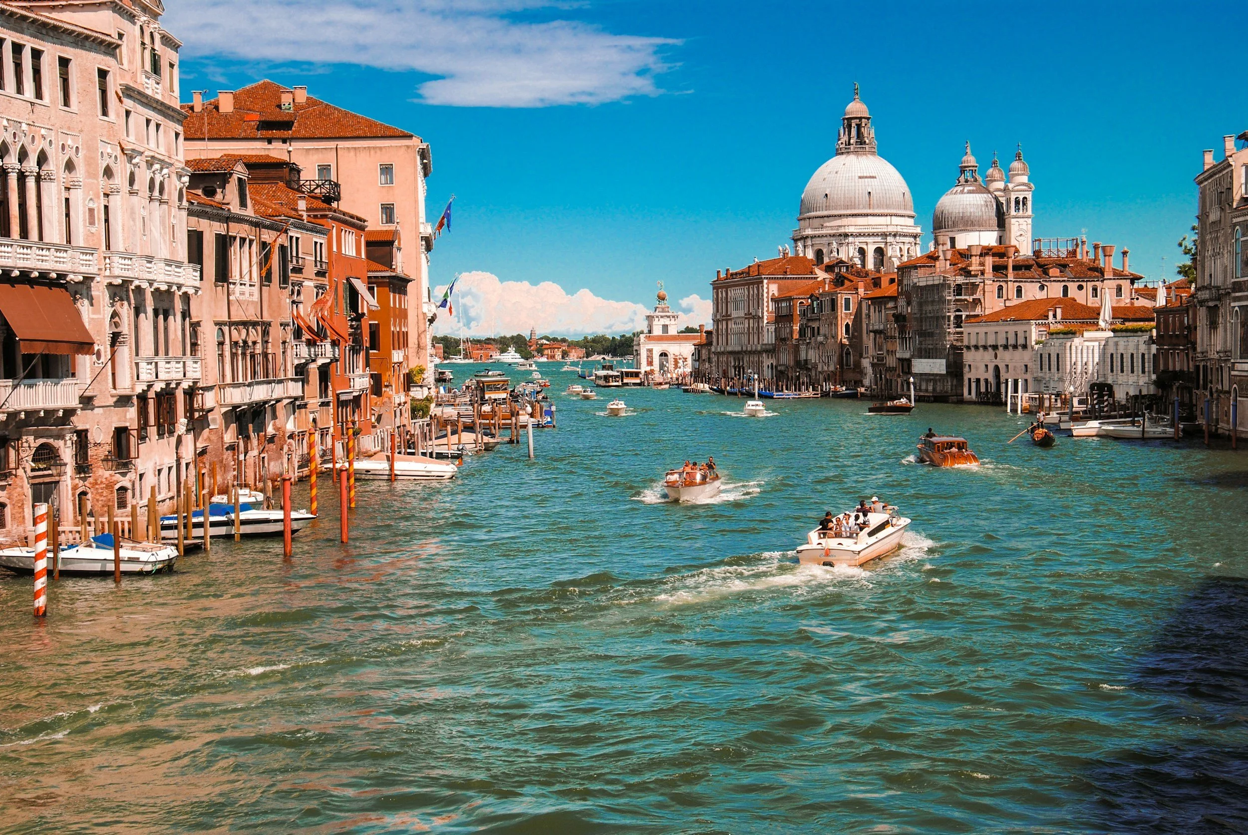 View of a canal in Venice, Italy, with boats and historic buildings along the water, and the dome of a church in the background under a partly cloudy sky.