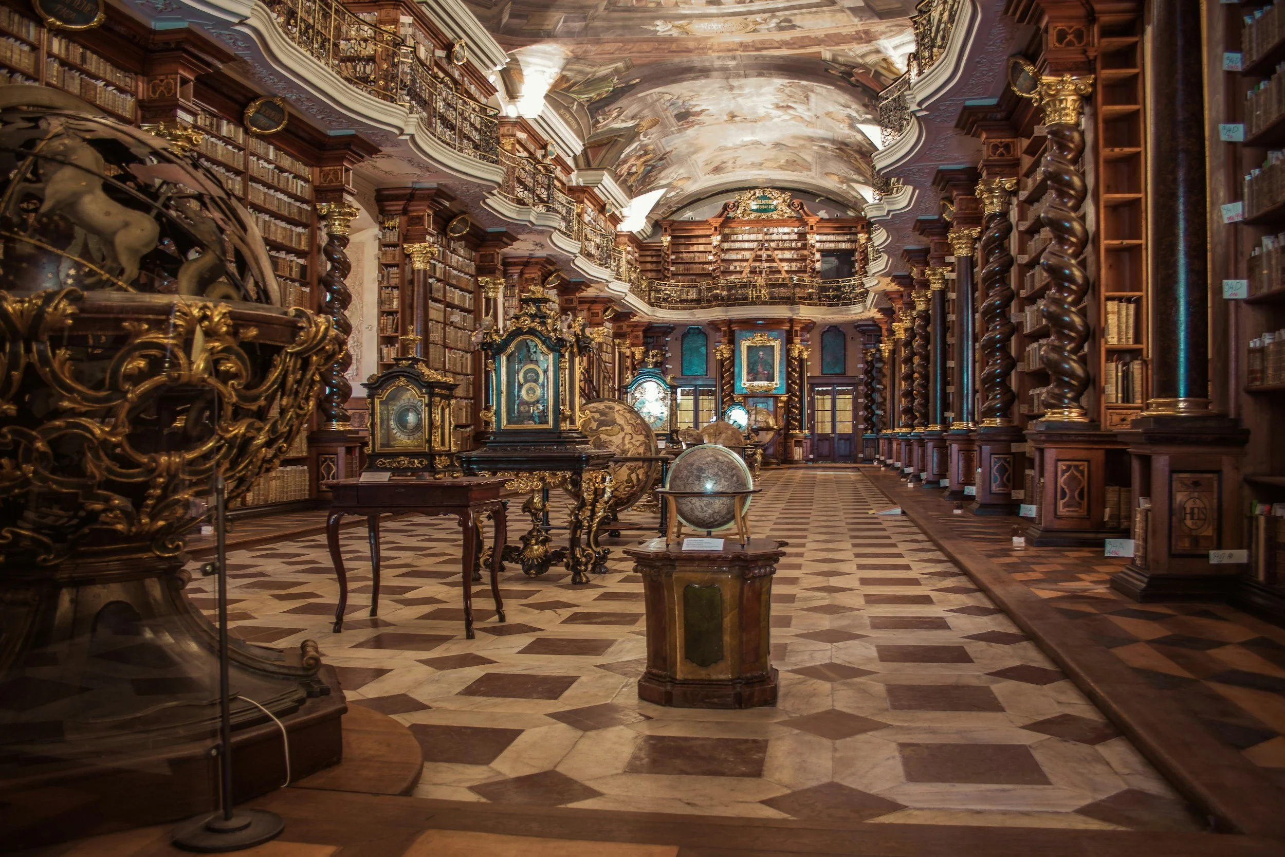 An elaborate, historical library with wooden bookshelves, globes, and ornate decorations, including gold accents and a painted ceiling.