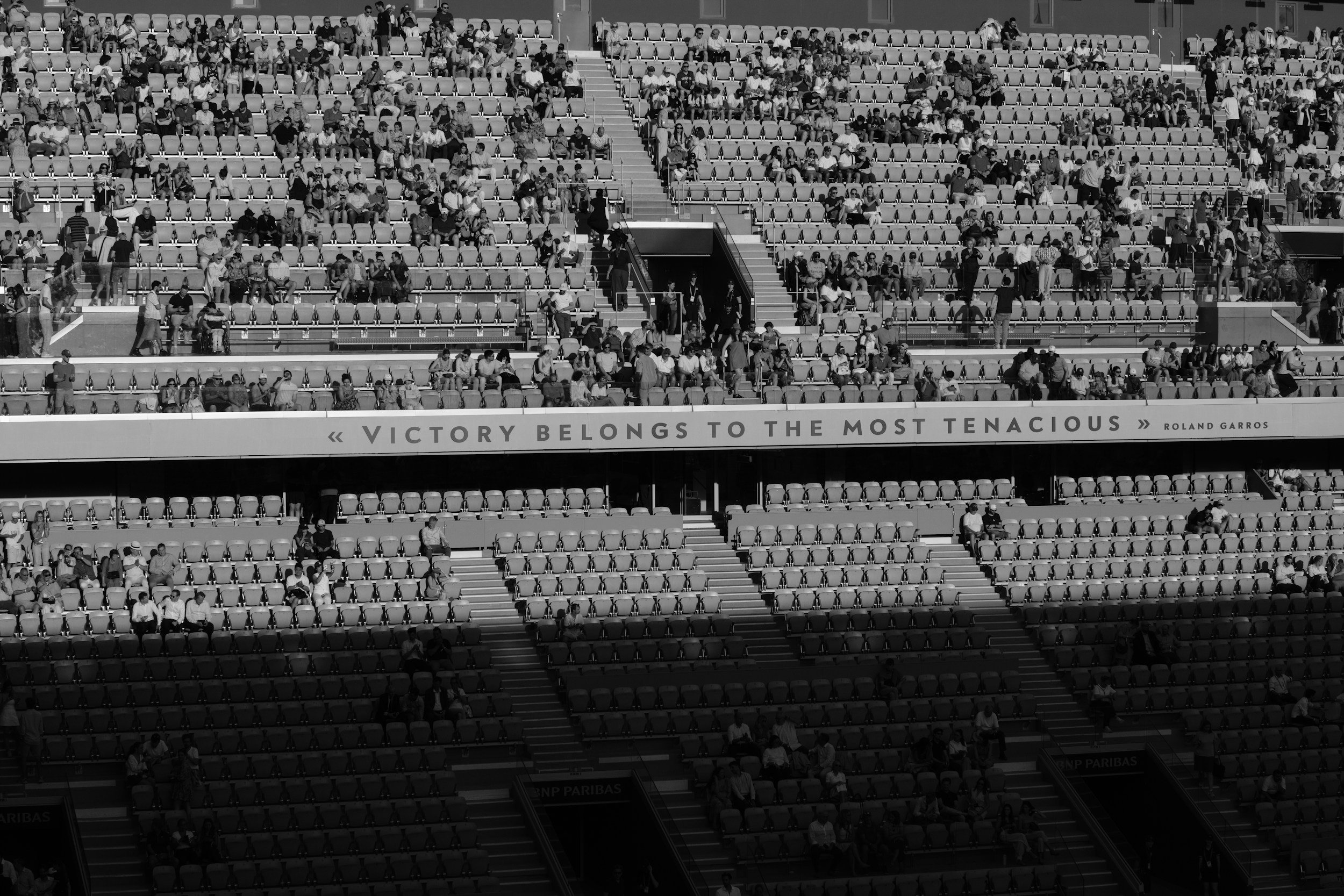 Crowded stadium seating with spectators, stairs in the middle, and a quote on the front reading "Victory belongs to the most tenacious" by Roland Garros.
