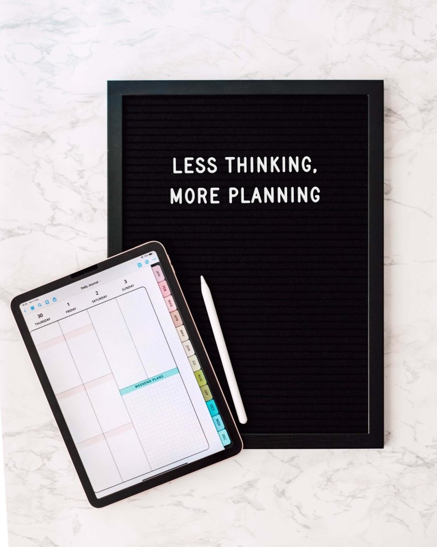 A tablet displaying a weekly planner, a white stylus, and a black message board with the text 'Less thinking, more planning' written on it, all on a white marble surface.