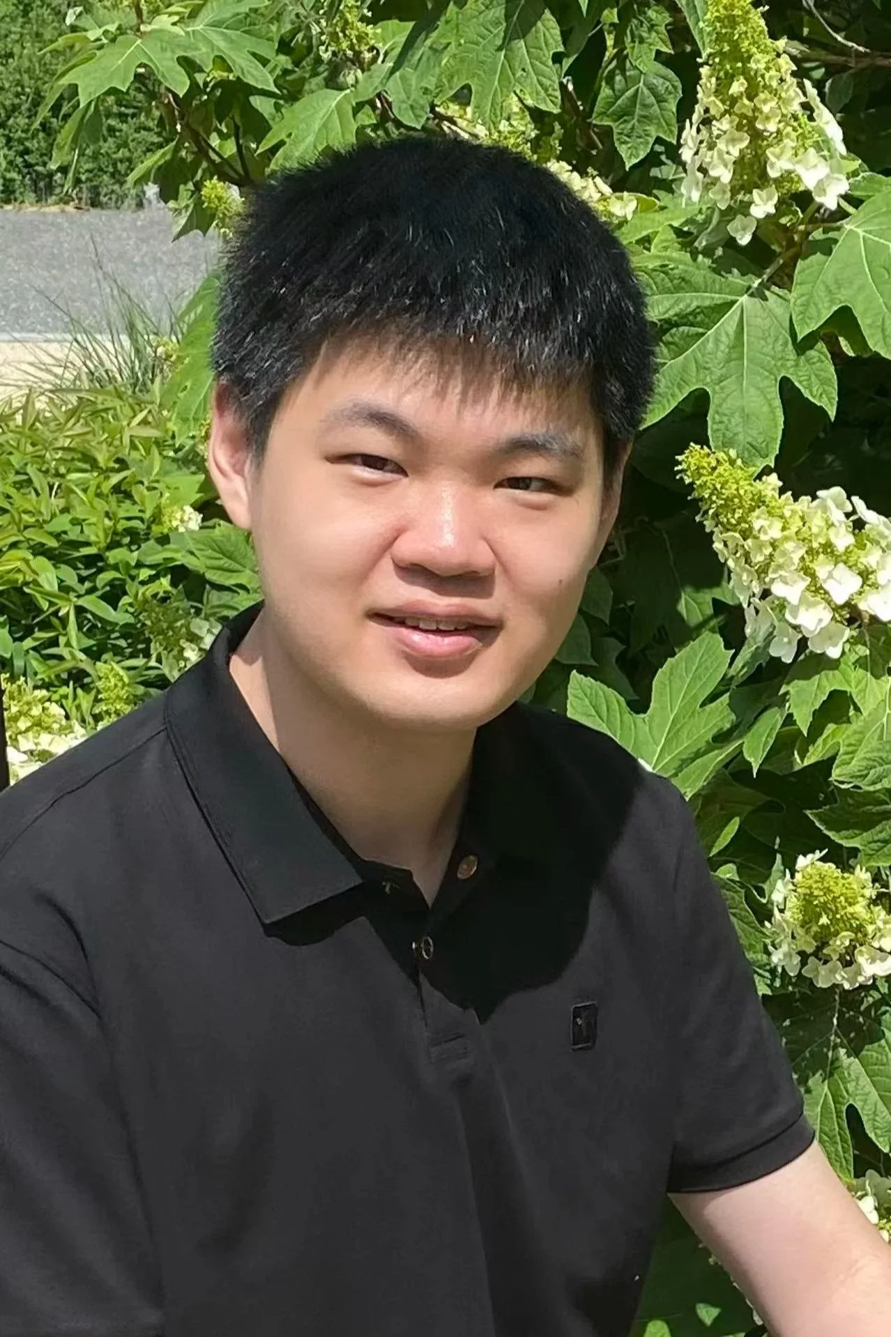 A young man with short black hair and fair skin, wearing a black polo shirt, sitting outdoors in front of lush green foliage with white flowers.