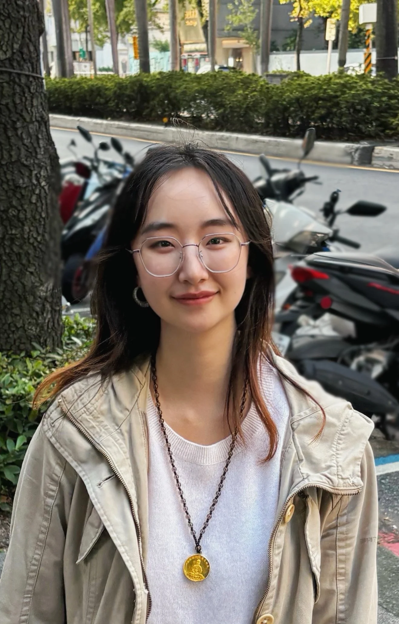 A young woman with glasses, earrings, and shoulder-length hair, smiling outdoors with parked motorcycles and a city street in the background.