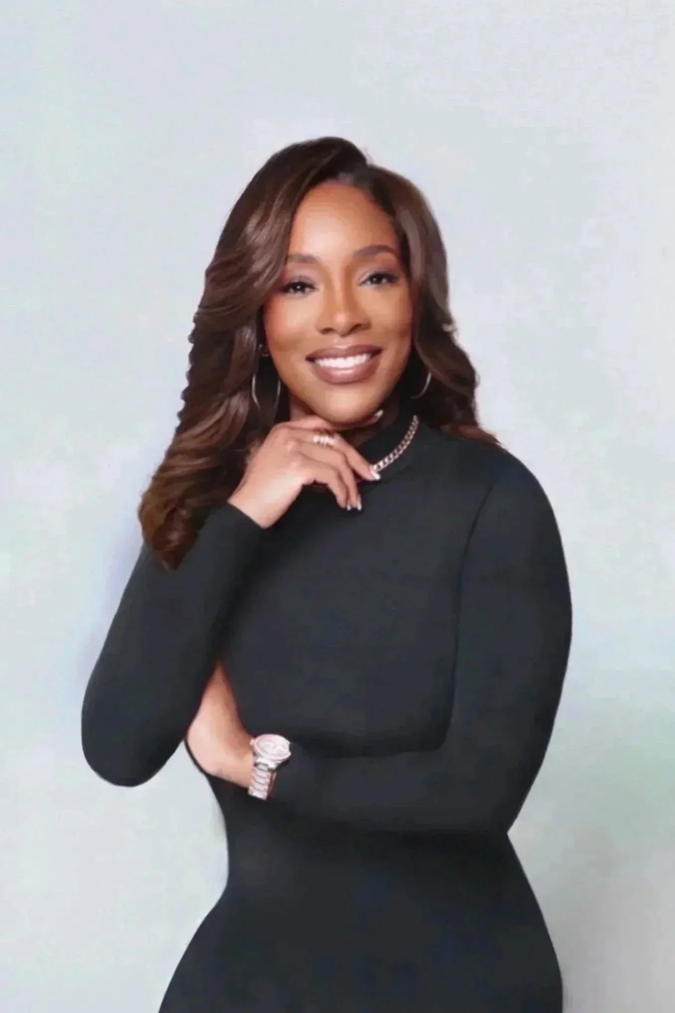 A woman with long wavy hair dressed in black, smiling and posing with her hand near her chin against a light-colored background.